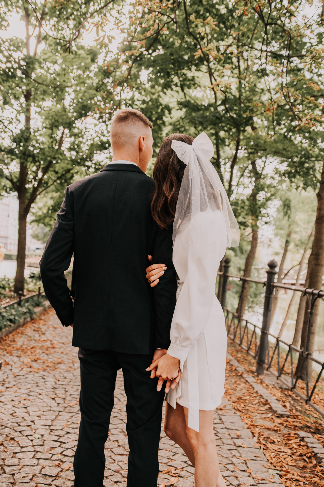 Para młoda, widok z tyłu, trzymająca się za ręce na kamiennej ścieżce w parku. Panna młoda w sukni z kokardą we włosach, pan młody w czarnym garniturze.