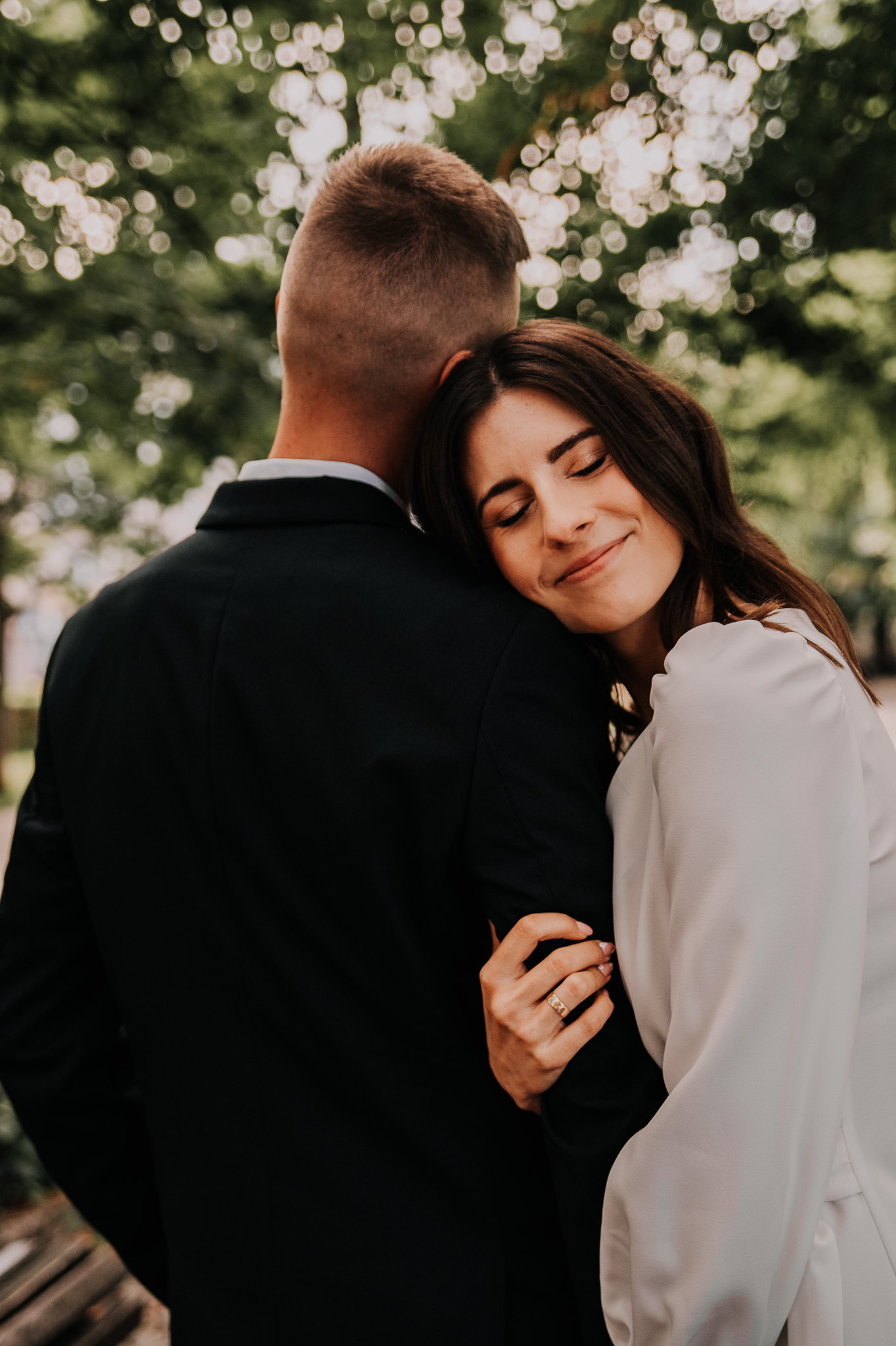 Para w objęciach na tle rozmytej zieleni. Kobieta z zamkniętymi oczami opiera głowę o ramię mężczyzny. Widoczny złoty pierścionek na jej palcu.