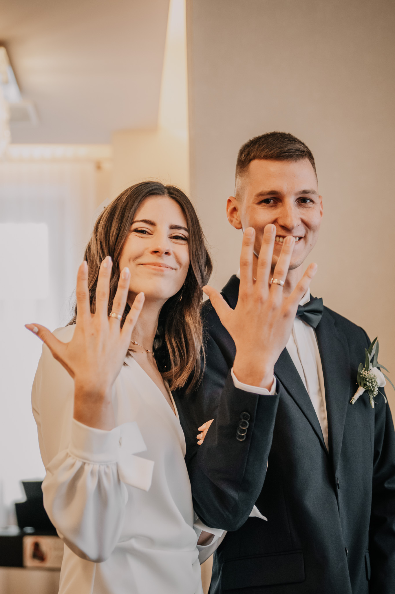 Uśmiechnięta para młoda prezentuje obrączki ślubne na dłoniach, on w czarnym smokingu z butonierką, ona w białej sukience, jasne wnętrze w tle.