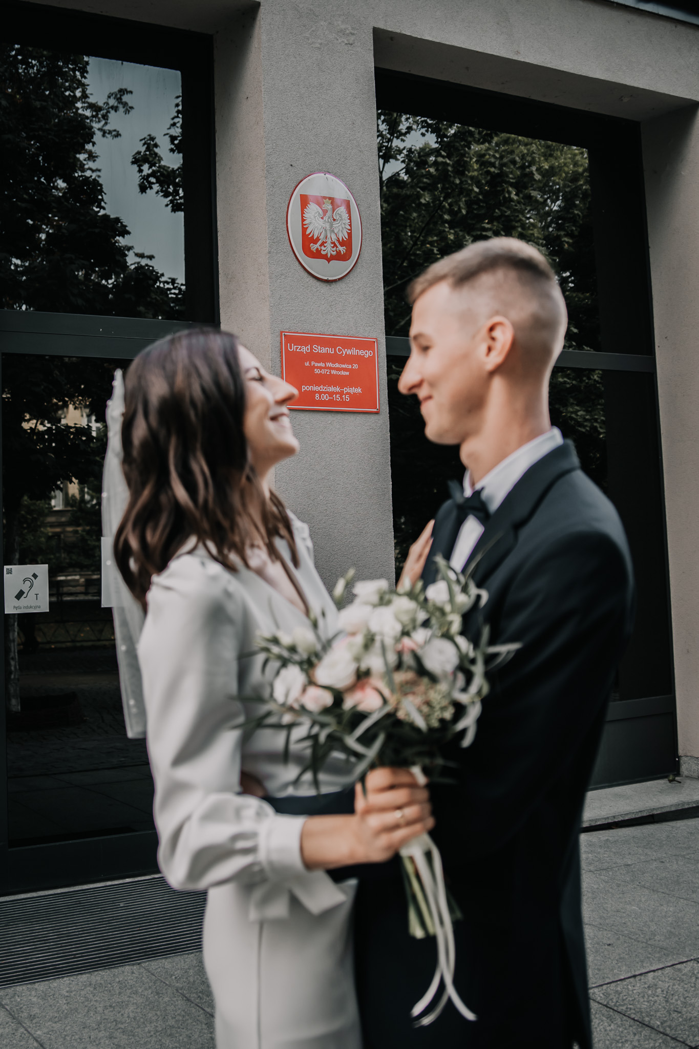 Uśmiechnięta para młoda przed Urzędem Stanu Cywilnego we Wrocławiu. Panna młoda w białej sukni trzyma bukiet kwiatów, pan młody w czarnym garniturze.