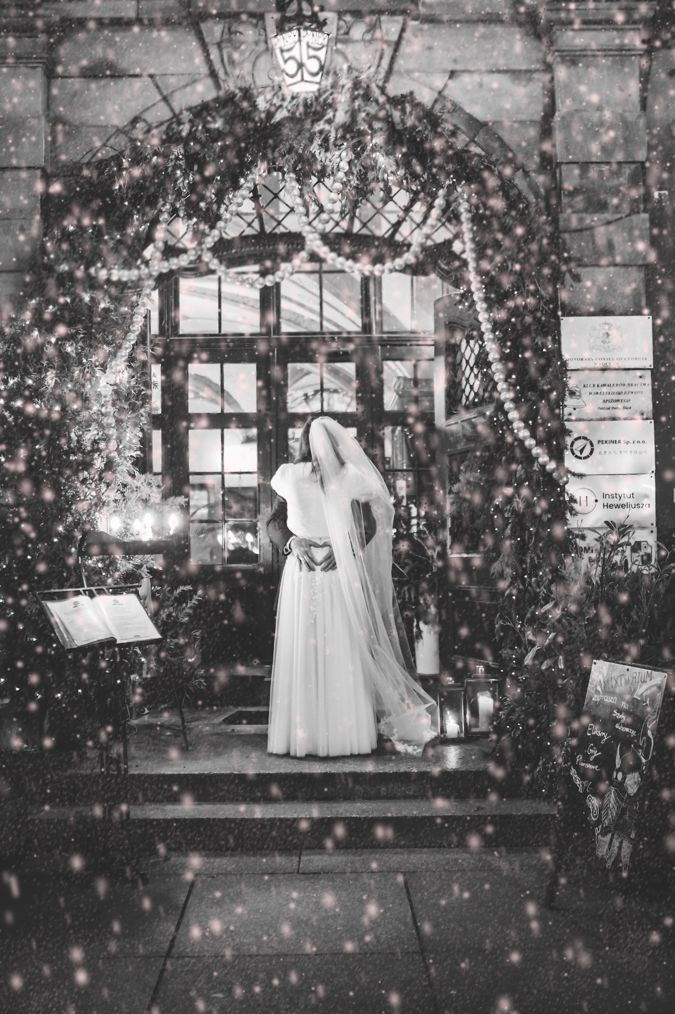 Para młoda w objęciach przed wejściem do budynku, dekoracje świąteczne, czarno-białe zdjęcie z efektem śniegu, widoczne lampiony i tablica informacyjna.