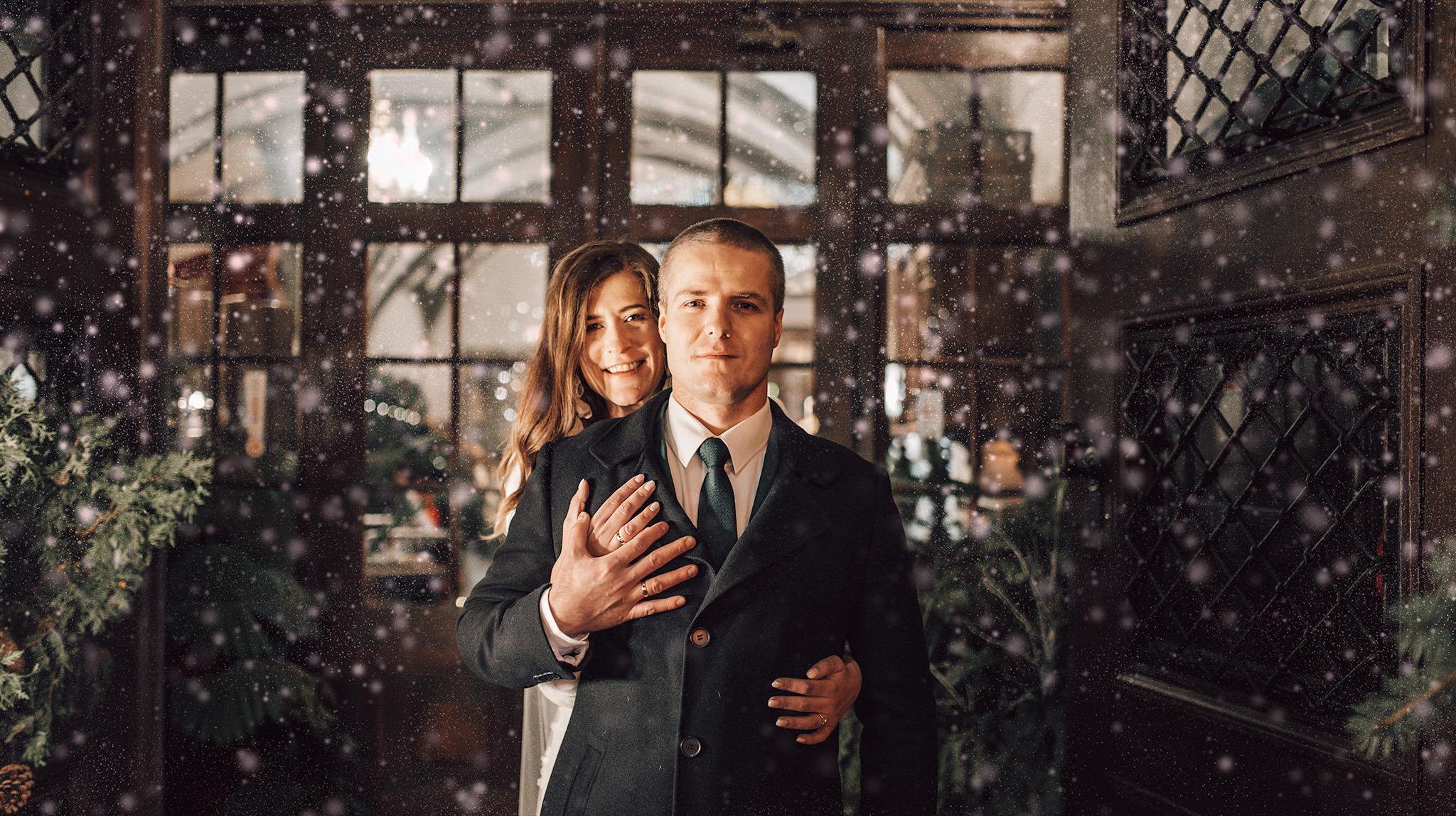 Uśmiechnięta para w zimowej scenerii, kobieta obejmuje mężczyznę. Efekt padającego śniegu, ciemne drewniane drzwi z okiennicami w tle, dekoracje świąteczne.