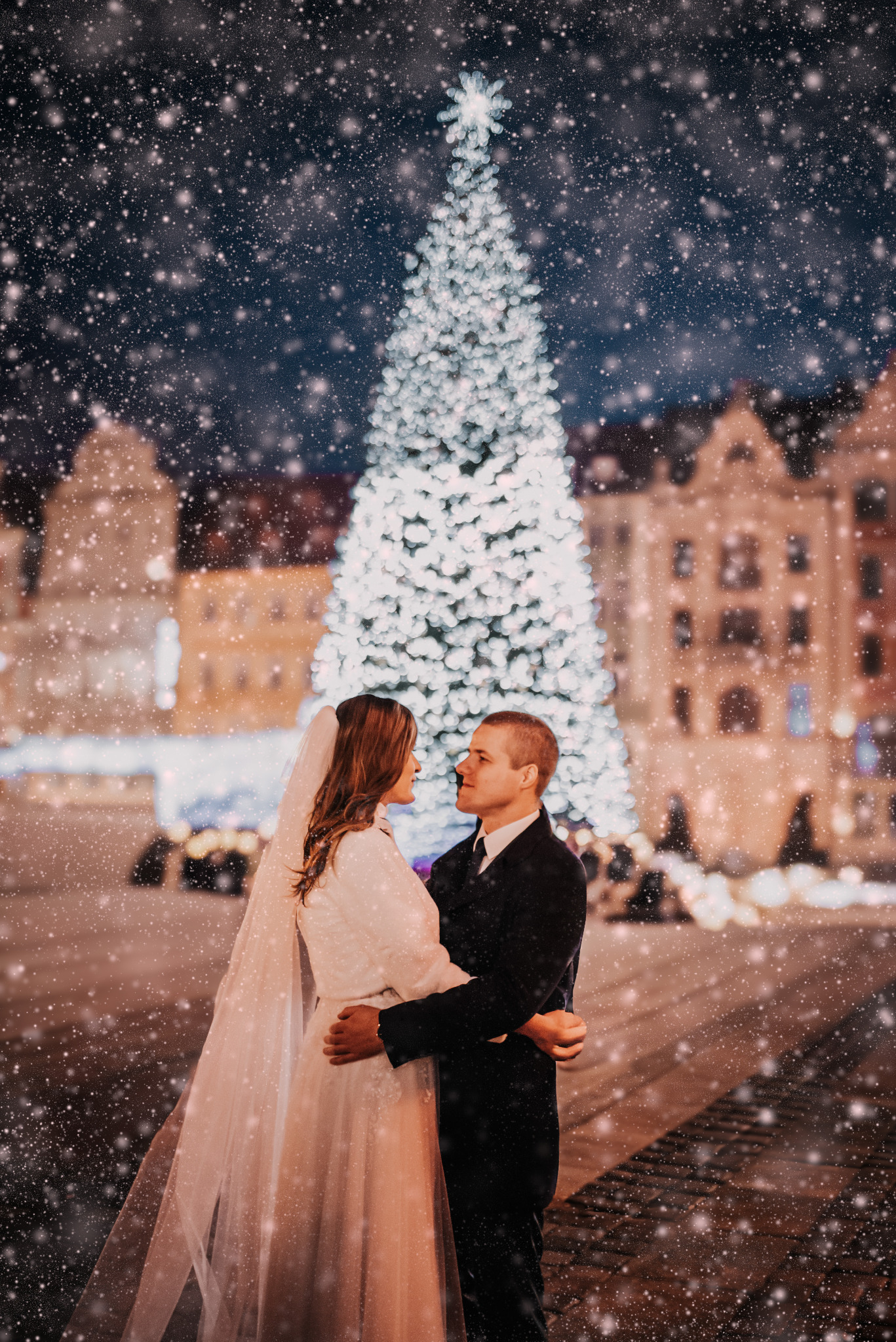 Zimowy ślub: para młoda w objęciach na tle rozświetlonej choinki i padającego śniegu na tle staromiejskiej architektury. Romantyczna atmosfera.