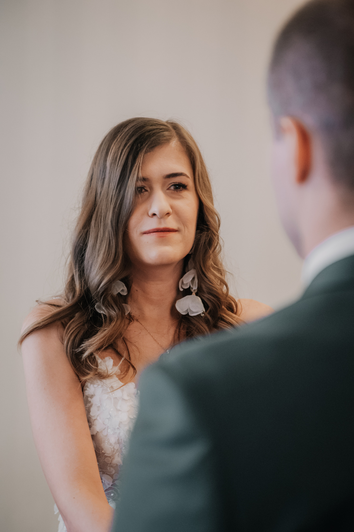 Panna młoda w sukni z aplikacjami kwiatowymi i dużymi kolczykami spogląda na pana młodego w ciemnozielonym garniturze podczas ceremonii ślubnej.