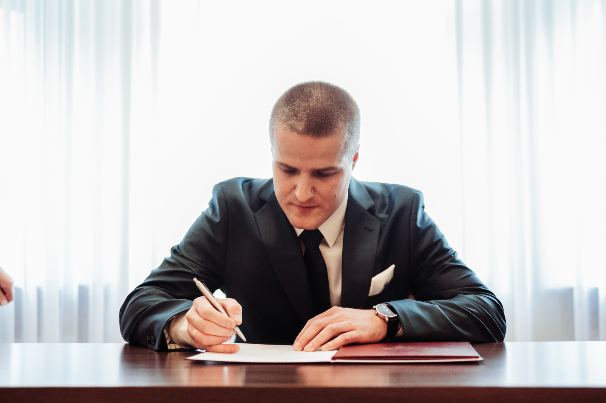 Mężczyzna w garniturze podpisuje dokument na drewnianym stole, w tle jasne zasłony. Widoczny zegarek na ręce i pióro w dłoni. Ujęcie z przodu, skupione na czynności podpisywania.
