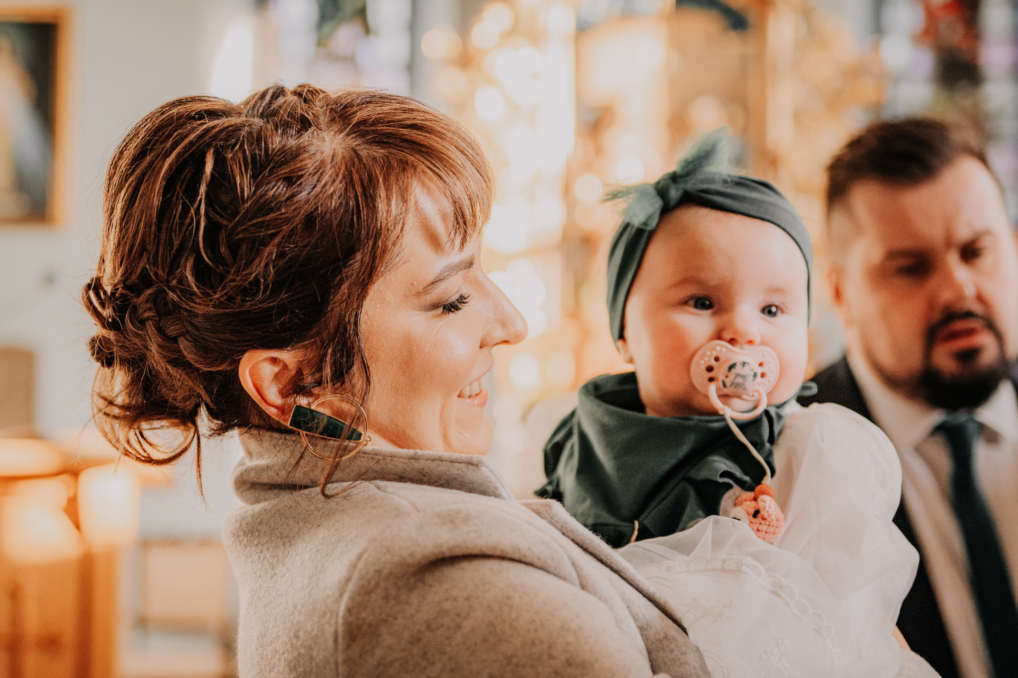 Uśmiechnięta kobieta trzyma na rękach niemowlę z smoczkiem, w tle mężczyzna. Ciepłe światło, portret rodzinny z uroczystości.