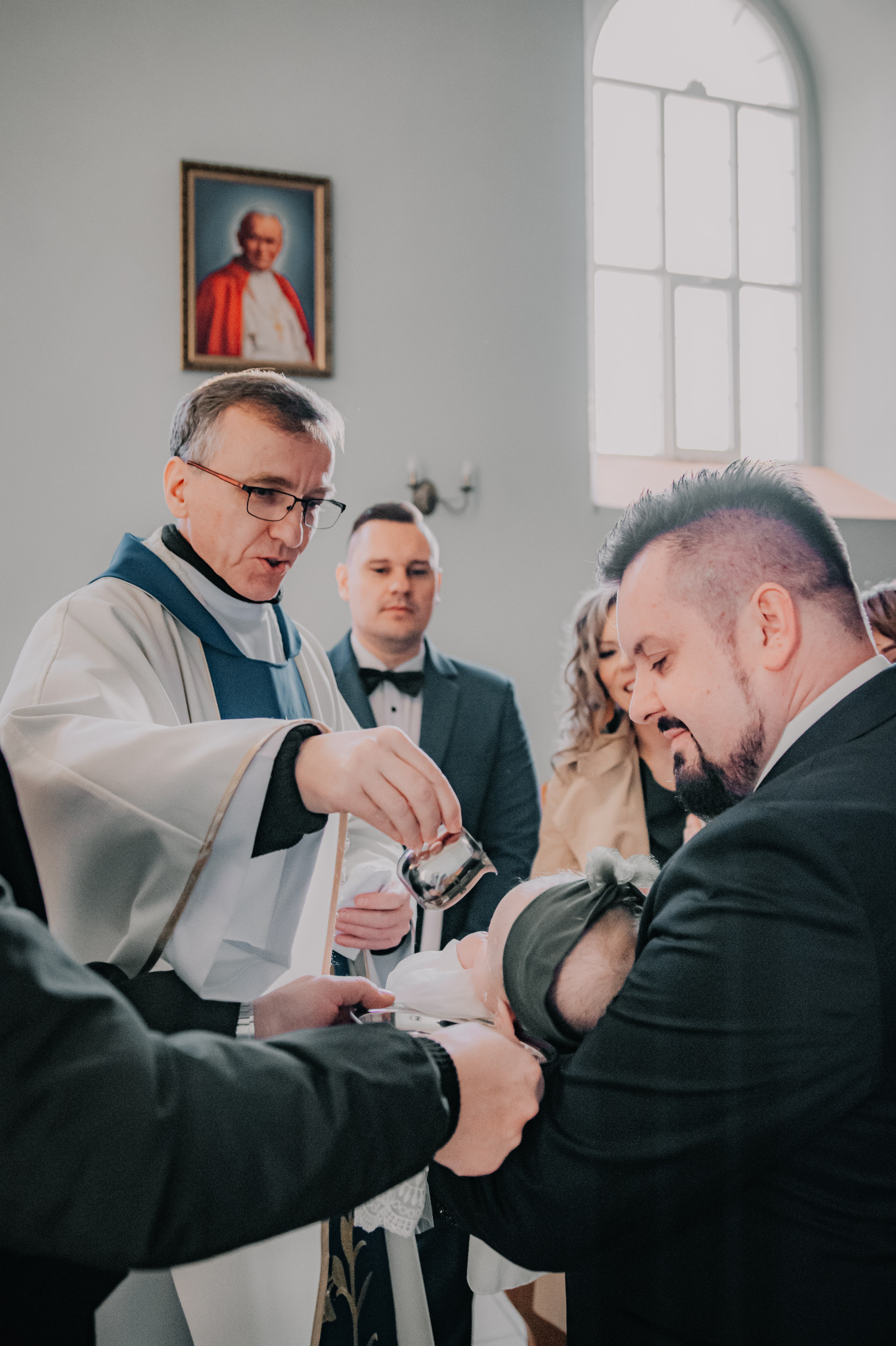 Chrzest dziecka w kościele: ksiądz polewa wodą główkę niemowlęcia trzymanego przez ojca. W tle rodzina i obraz Jana Pawła II.