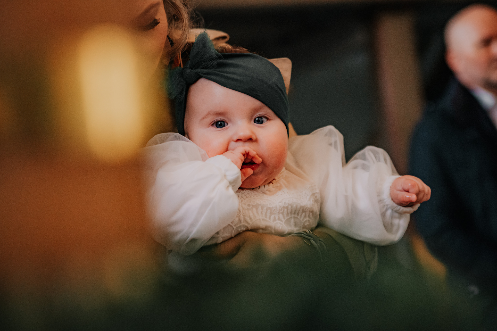 Urocze niemowlę w białej sukience i czarnej opasce, trzymane na rękach, z palcem w buzi. W tle rozmyte postacie, ciepłe oświetlenie, portret z uroczystości.