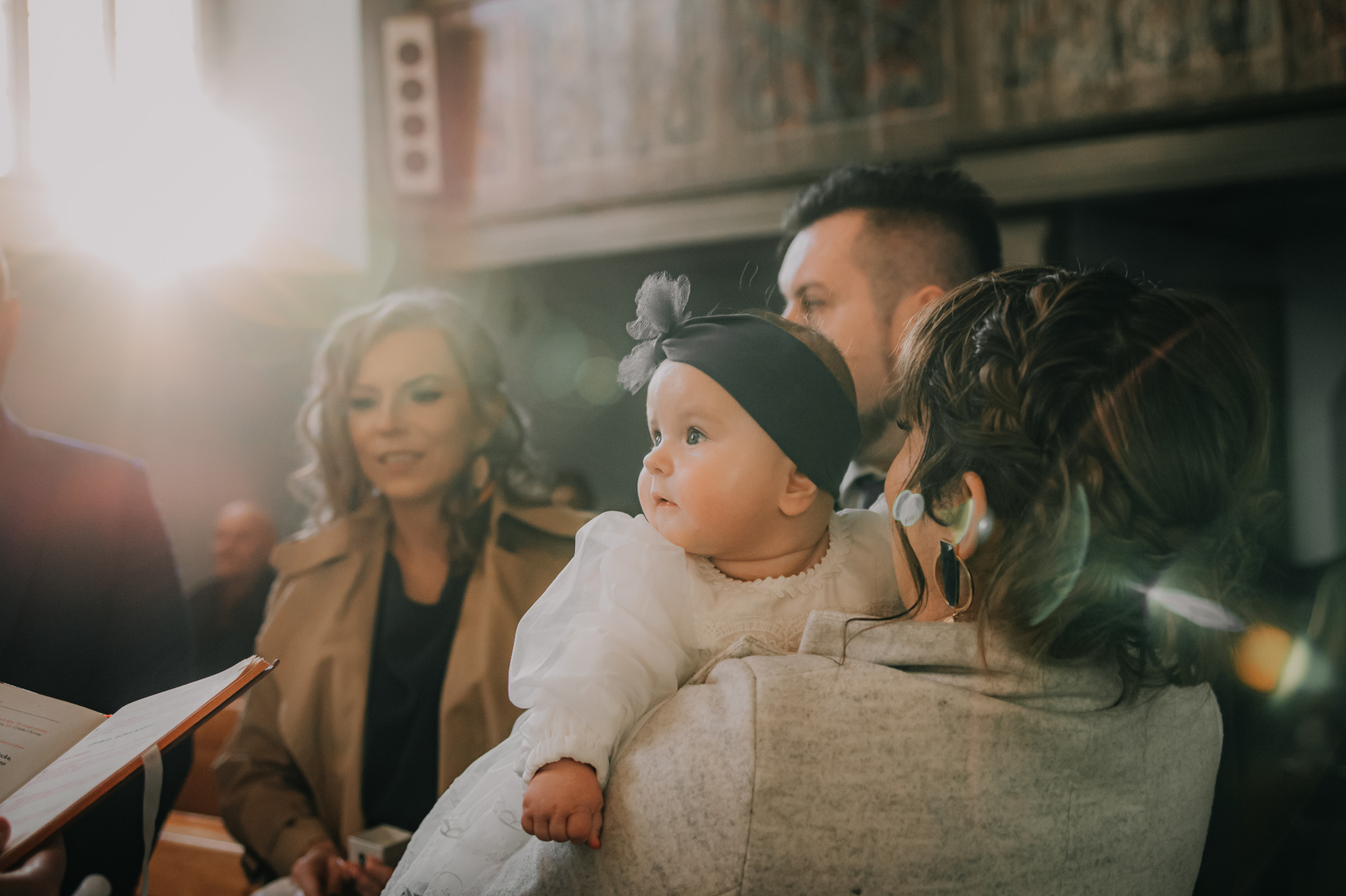 Ujęcie z uroczystości chrztu: matka trzyma na rękach ubraną w białą sukienkę dziewczynkę z czarną opaską, w tle widoczni goście i wnętrze kościoła.