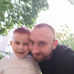 Uśmiechnięty mężczyzna z brodą i mały chłopiec z rudymi włosami pozują do zdjęcia na tle zieleni i budynku. Jasne, naturalne światło.