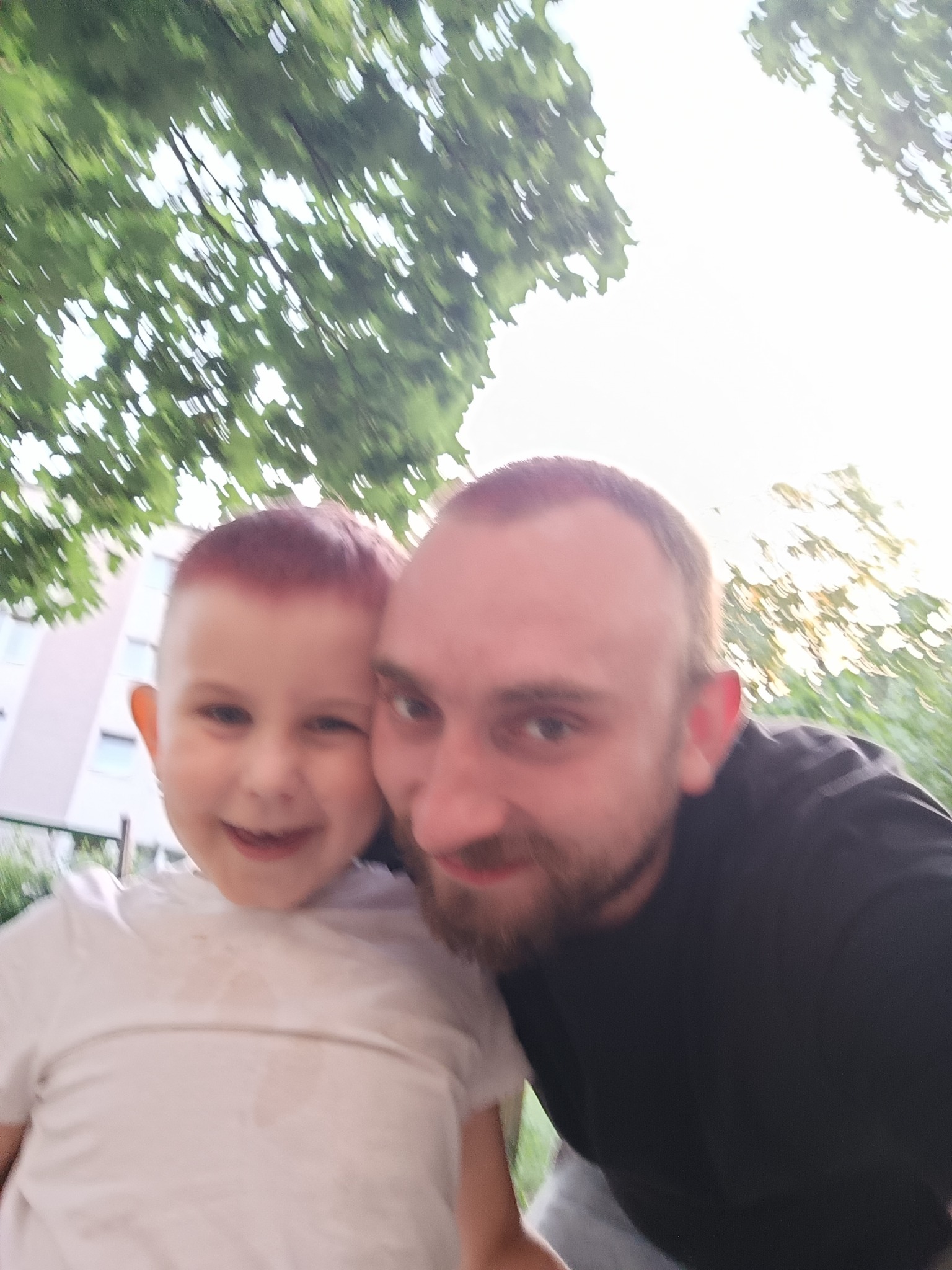 Uśmiechnięty mężczyzna z brodą i mały chłopiec z rudymi włosami pozują do zdjęcia na tle zieleni i budynku. Jasne, naturalne światło.