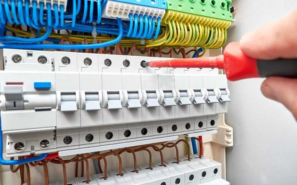 Elektryk dokręca śrubokrętem zabezpieczenia w rozdzielnicy elektrycznej. Widoczne kable, bezpieczniki i elementy instalacji.