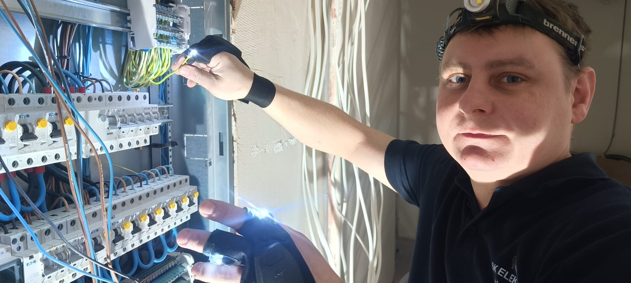 Elektryk w trakcie pracy nad rozdzielnicą elektryczną, używający rękawic z wbudowanymi LEDami i latarki czołowej, w tle ściana z kablami.