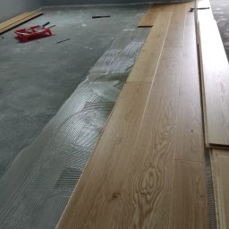 Volodymyr Rokush - Proces montażu jasnych paneli podłogowych, widoczny klej w charakterystyczne wzory, narzędzia i nieułożone deski w tle.