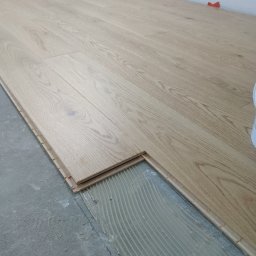 Volodymyr Rokush - Fragment podłogi z jasnych paneli drewnopodobnych w trakcie montażu, widoczna warstwa kleju i elementy dystansowe przy ścianie.