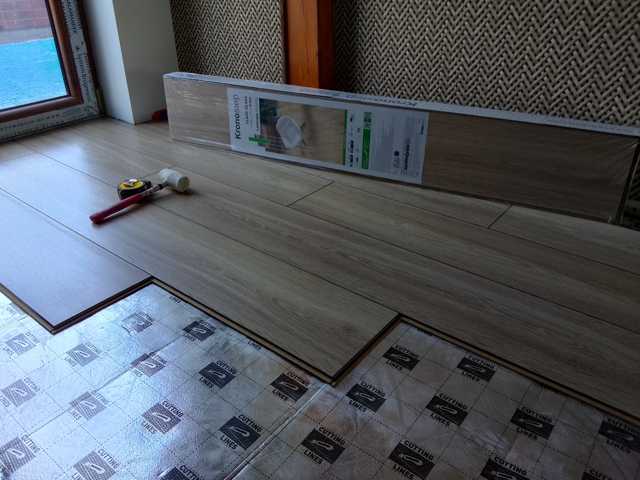 Początek prac: rozłożona folia podkładowa, częściowo ułożone panele podłogowe w odcieniach beżu, paczka paneli Kronostep i gumowy młotek obok okna.