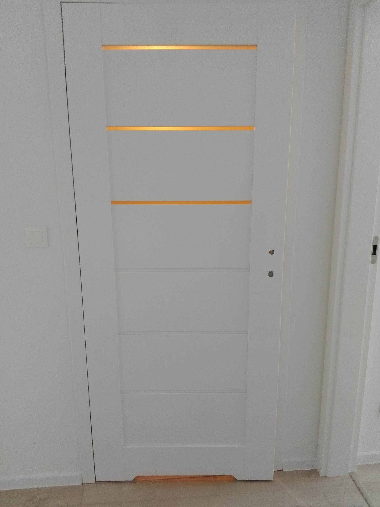 Białe drzwi wewnętrzne z poziomymi przeszkleniami, podświetlonymi od wewnątrz, zamontowane w białej ścianie.