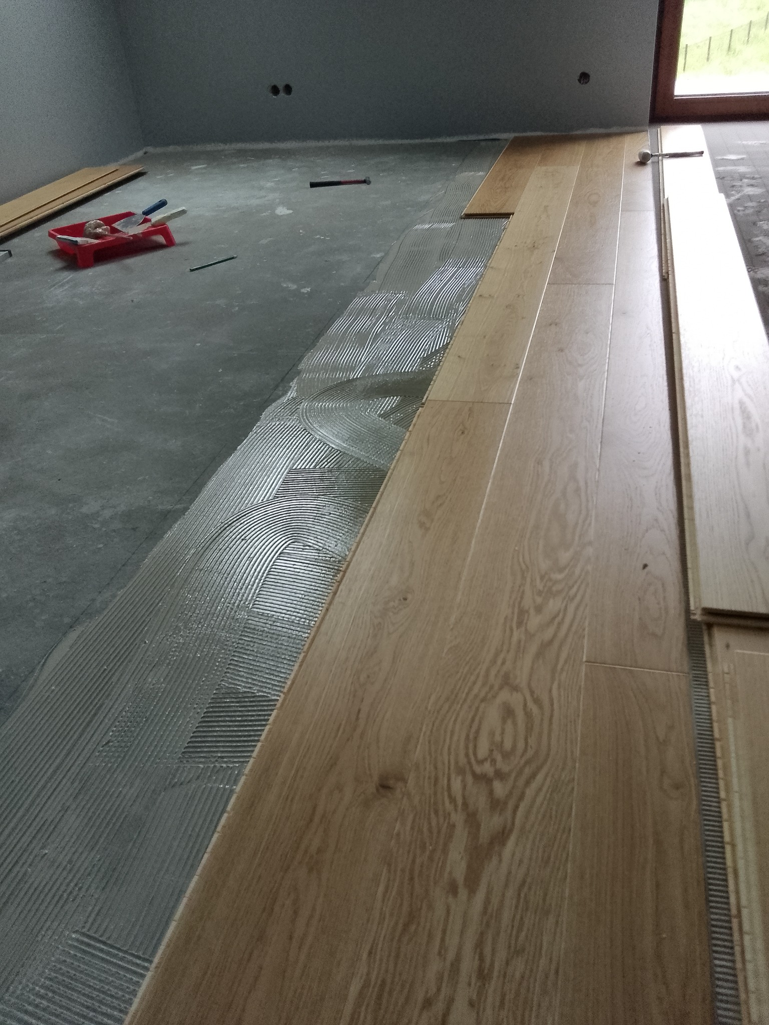 Proces montażu jasnych paneli podłogowych, widoczny klej w charakterystyczne wzory, narzędzia i nieułożone deski w tle.