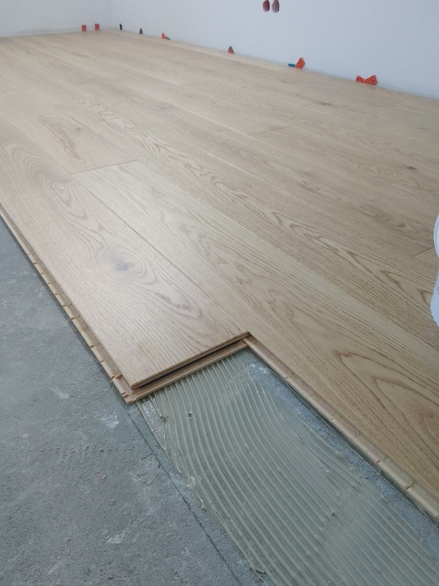 Fragment podłogi z jasnych paneli drewnopodobnych w trakcie montażu, widoczna warstwa kleju i elementy dystansowe przy ścianie.