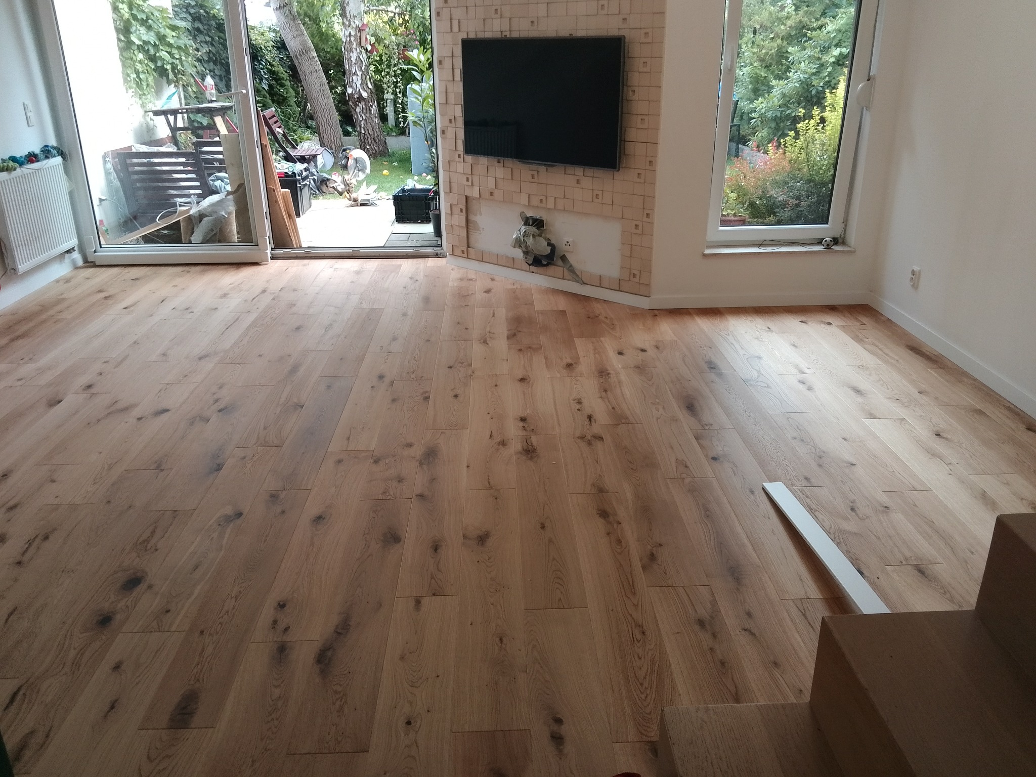 Ułożona podłoga z drewnianych desek w jasnym odcieniu dębu, widoczne schody, listwa przypodłogowa i ściana z telewizorem w tle.