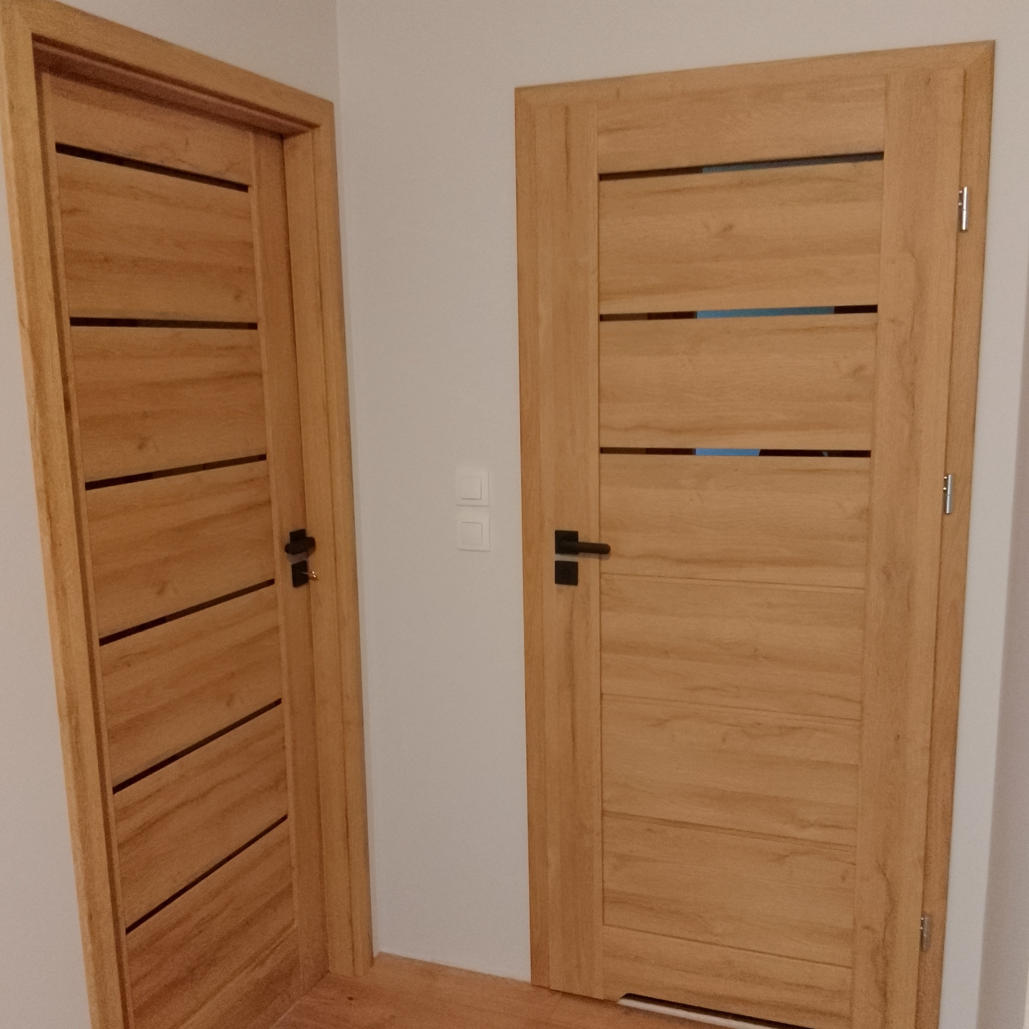 Dwie drewniane drzwi wewnętrzne z poziomymi wstawkami, czarne klamki, widoczne na tle białej ściany z włącznikiem.