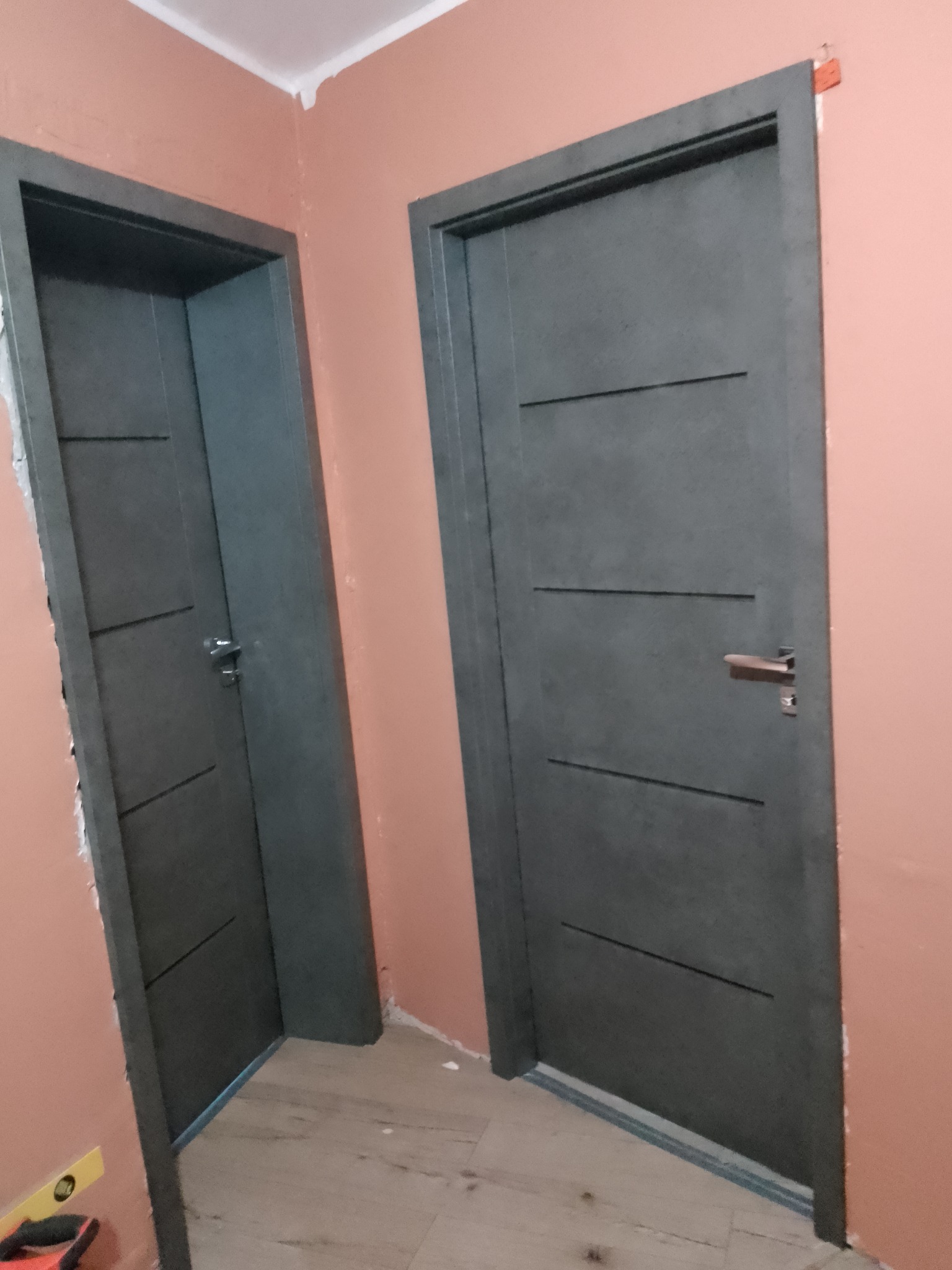 Montaż dwóch szarych drzwi wewnętrznych z poziomymi frezowaniami w ościeżnicach, widoczne ślady pianki montażowej na ścianie, poziomica przy jednej z ościeżnic.