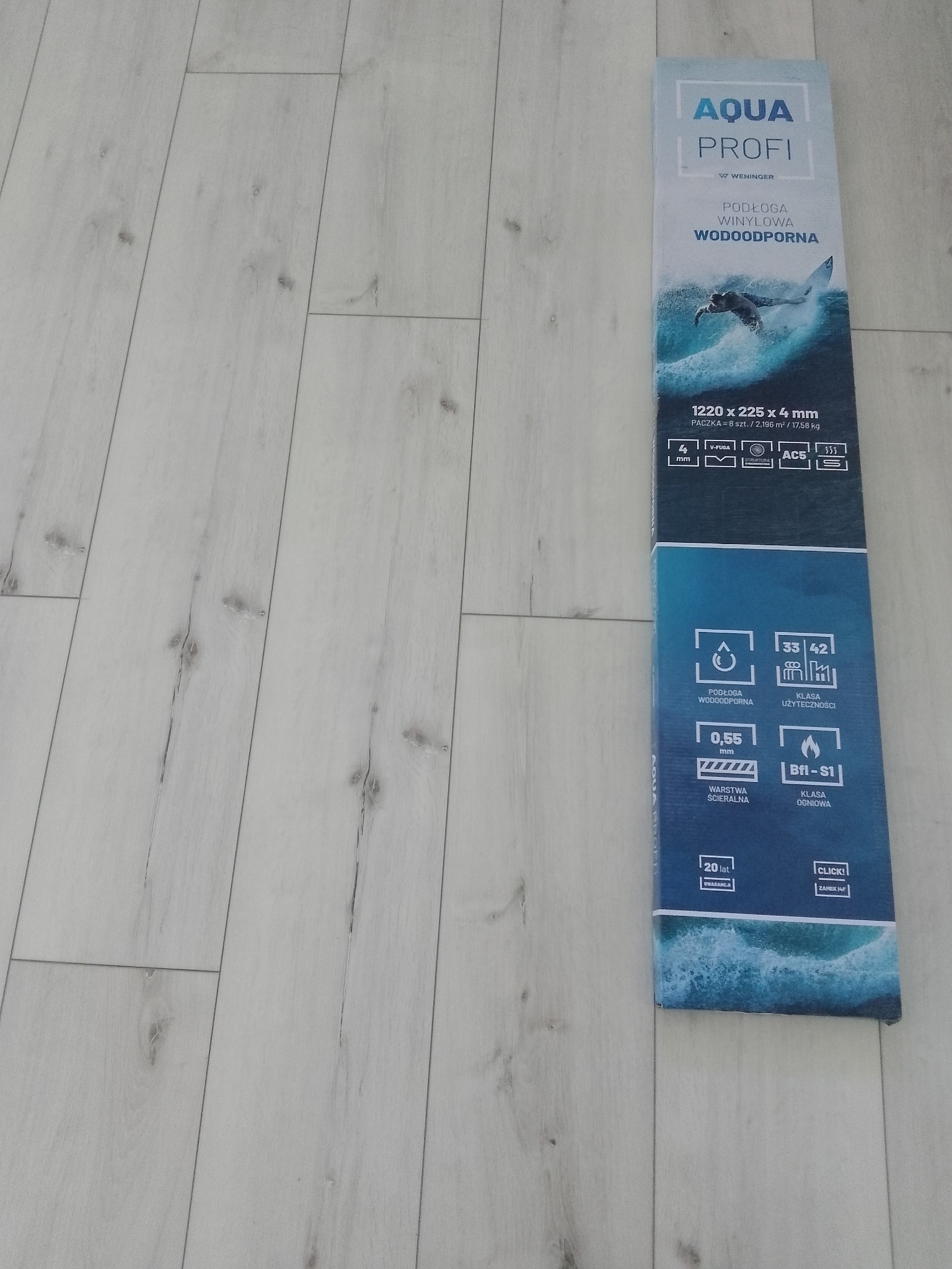 Opakowanie paneli winylowych wodoodpornych Aqua Profi na tle ułożonych paneli o jasnym, drewnopodobnym wzorze.