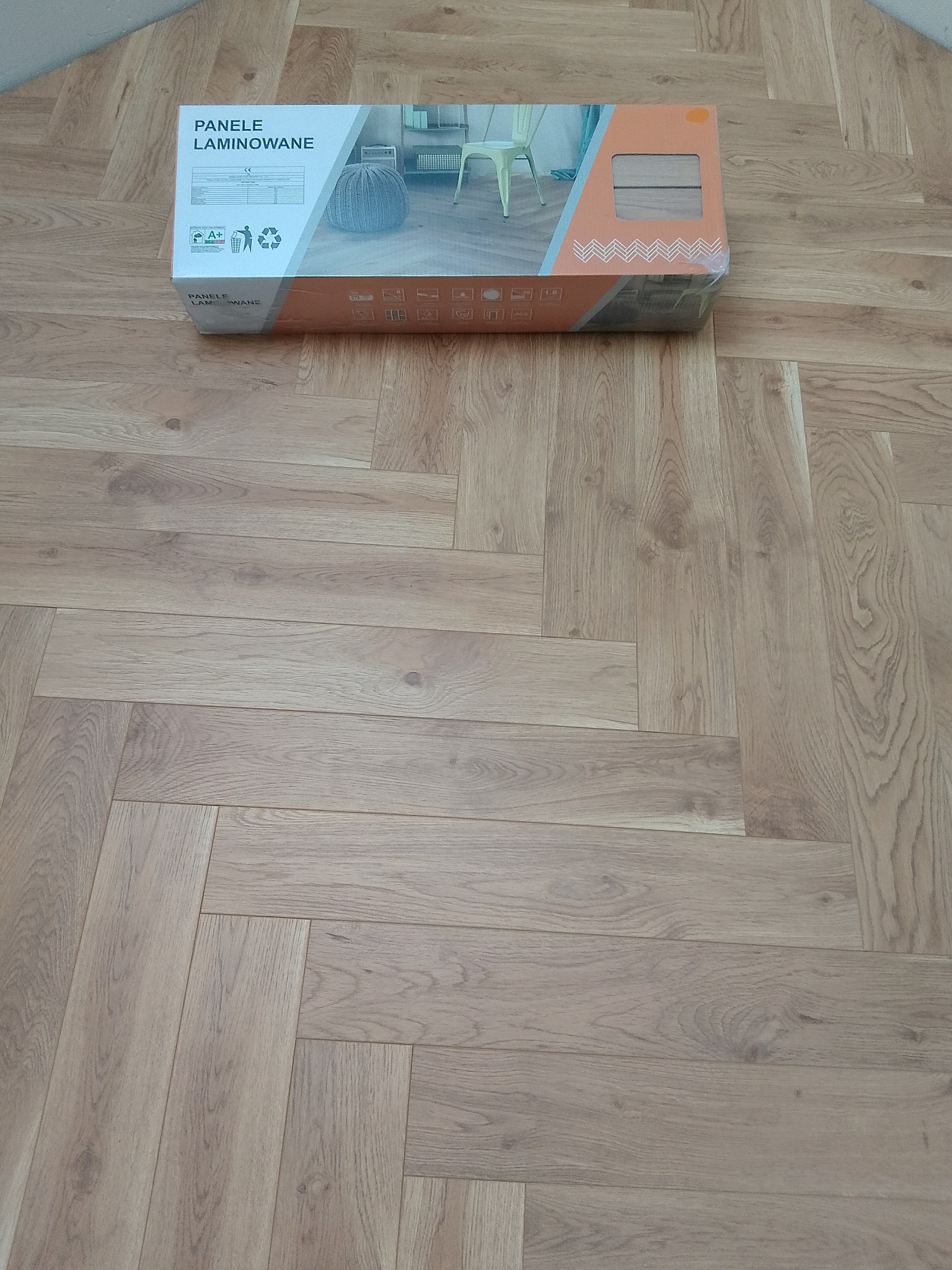 Ułożone panele laminowane w jodełkę, obok leży karton z tymi samymi panelami