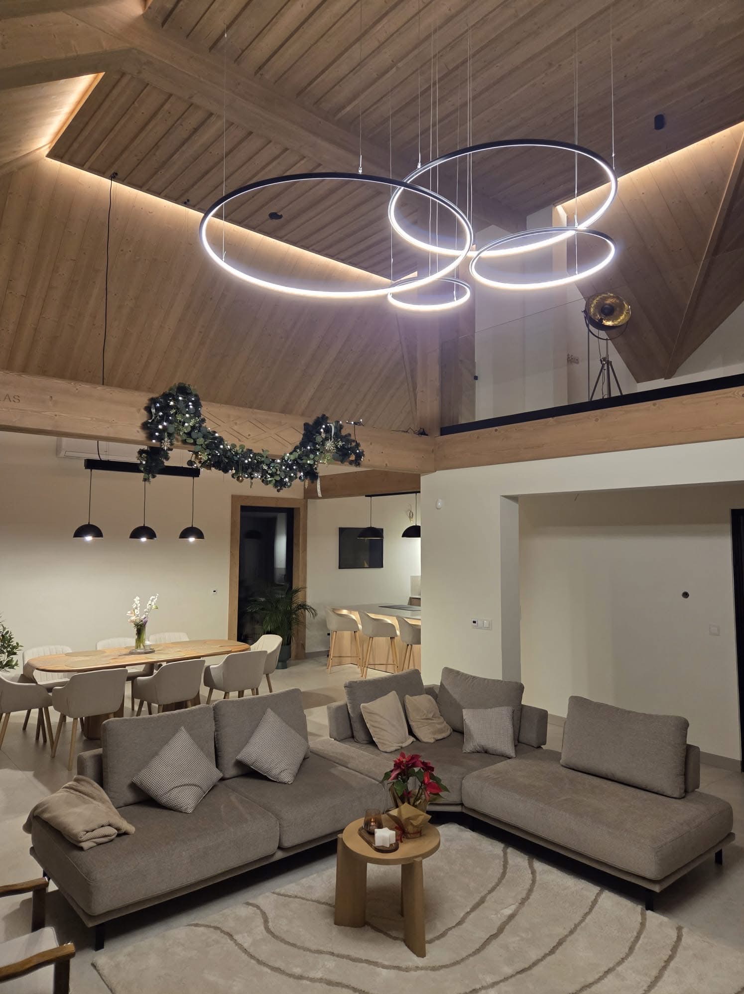 Nowoczesne oświetlenie w przestronnym salonie z drewnianym stropem. Okrągłe lampy wiszące, minimalistyczny design, ciepłe światło. Widok na jadalnię i antresolę.