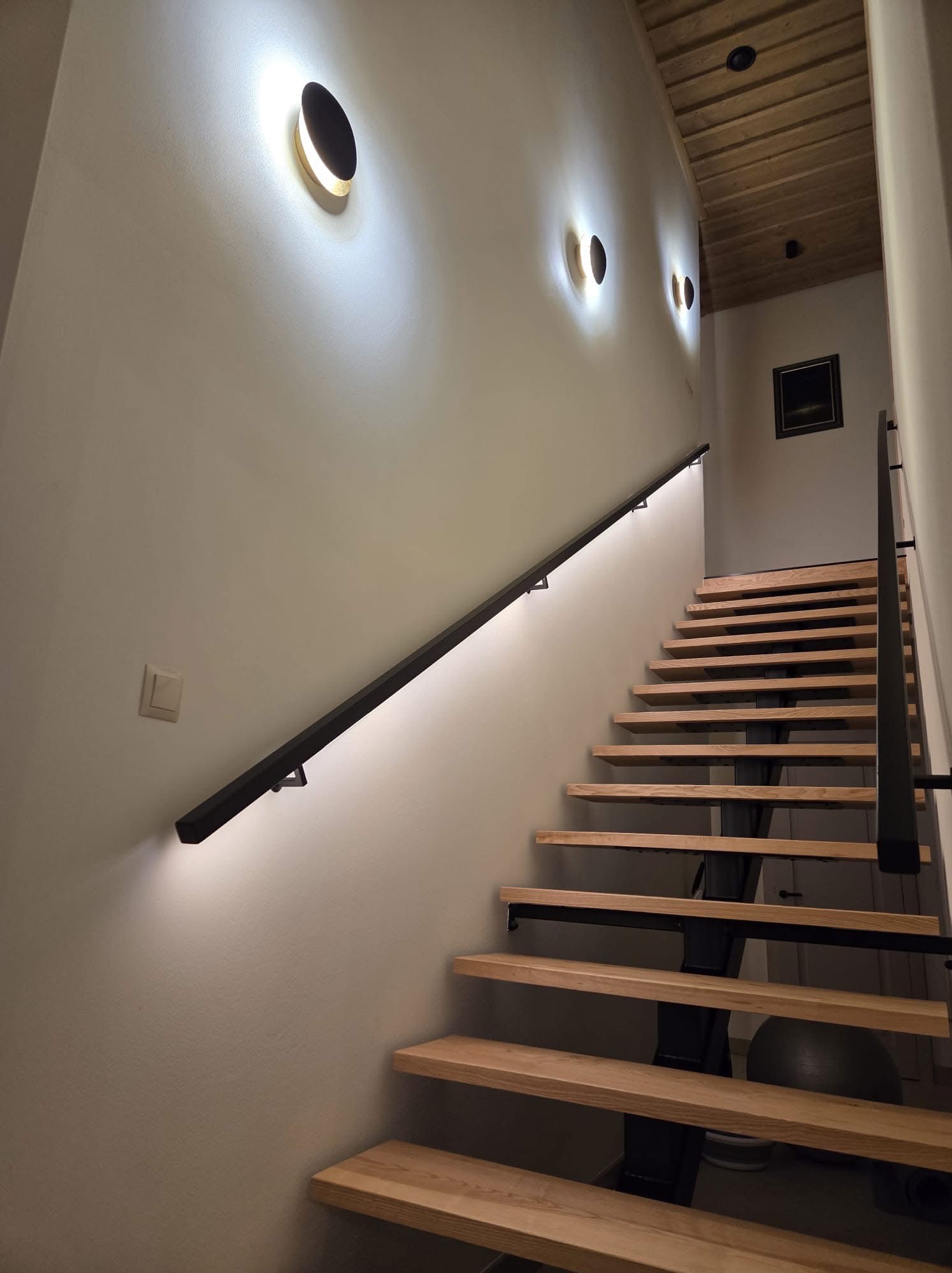 Schody z oświetleniem LED wzdłuż poręczy i okrągłe lampy ścienne w Nowym Targu. Minimalistyczny design, ciepłe światło.