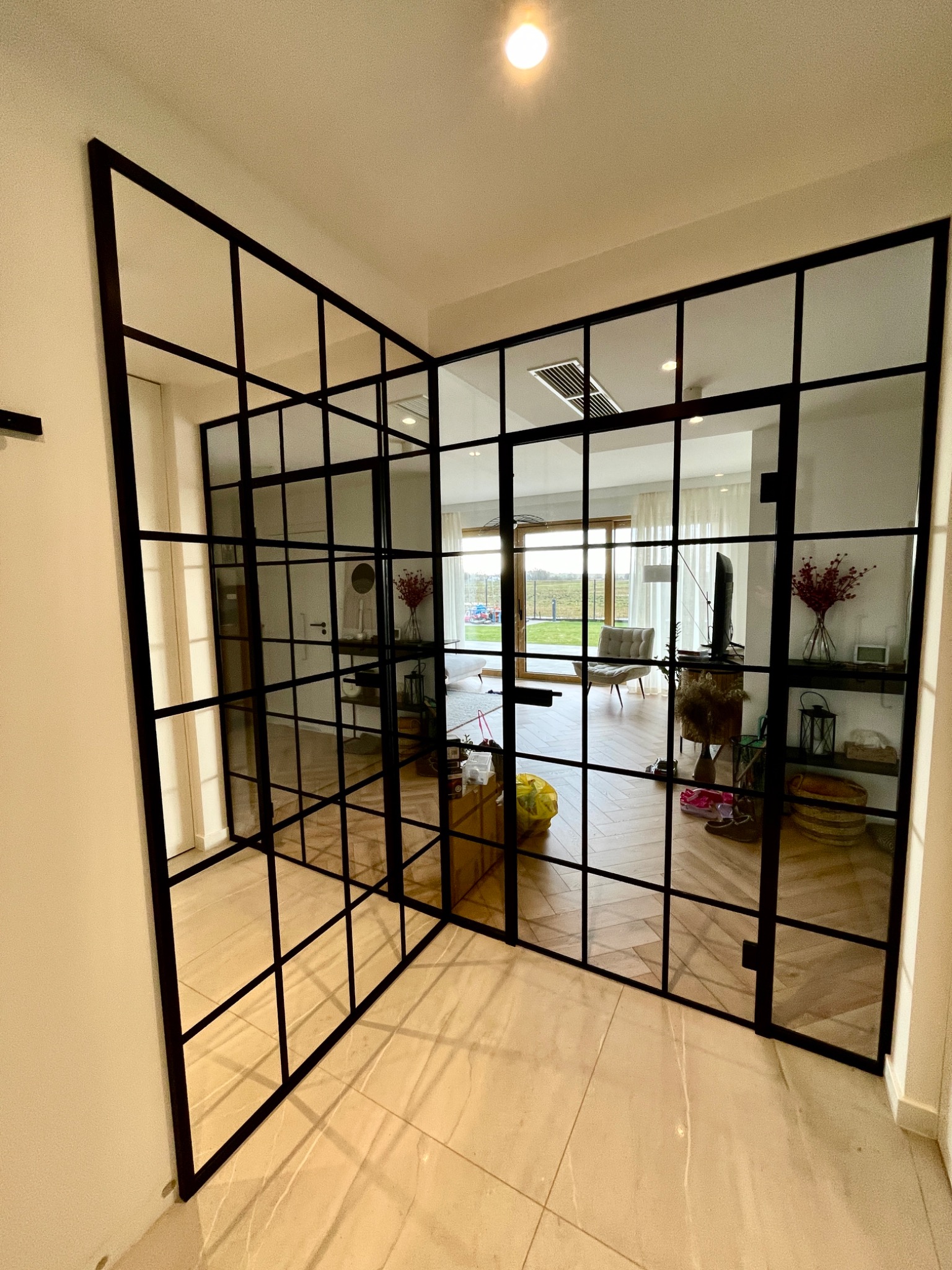 Nowoczesne, dwuskrzydłowe drzwi szklane z czarnymi szprosami oddzielające przedpokój od salonu, widoczne jasne płytki podłogowe i częściowo umeblowane wnętrze.