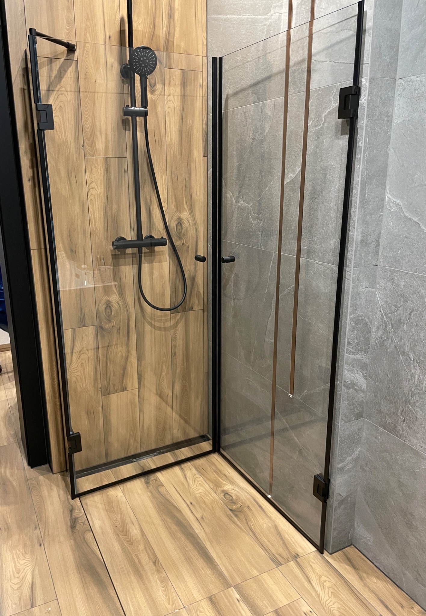 Szklana kabina prysznicowa z czarnymi profilami, drewnopodobne płytki na ścianie i podłodze, szara ściana z płytek imitujących kamień