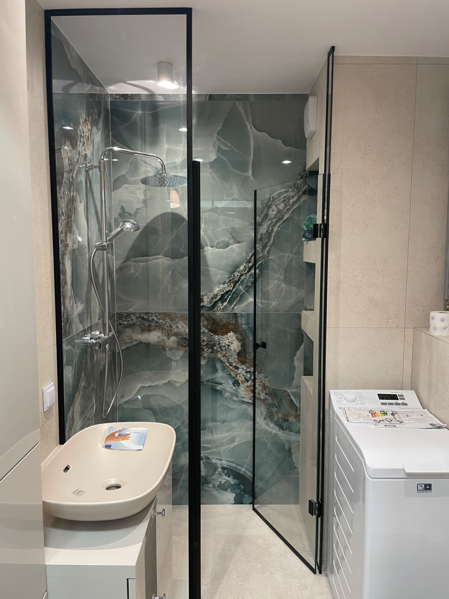 Nowoczesna kabina prysznicowa z czarnymi profilami i imitacją marmuru na ścianach, widoczna umywalka nablatowa i pralka.