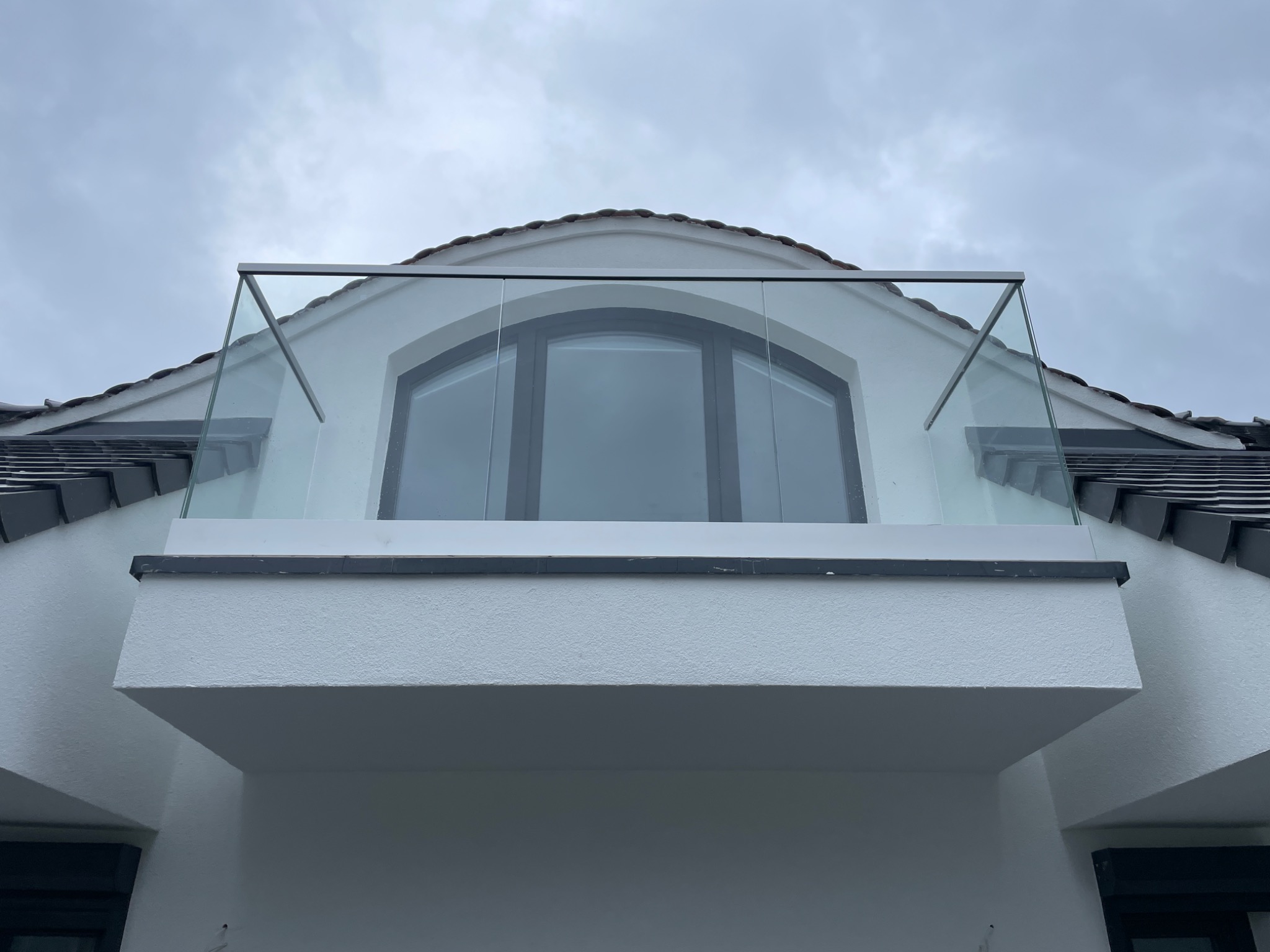 Zewnętrzny balkon z balustradą szklaną, widok z dołu na białą fasadę budynku z łukowym oknem i szklaną balustradą z metalowymi elementami mocującymi, pochmurne niebo w tle.