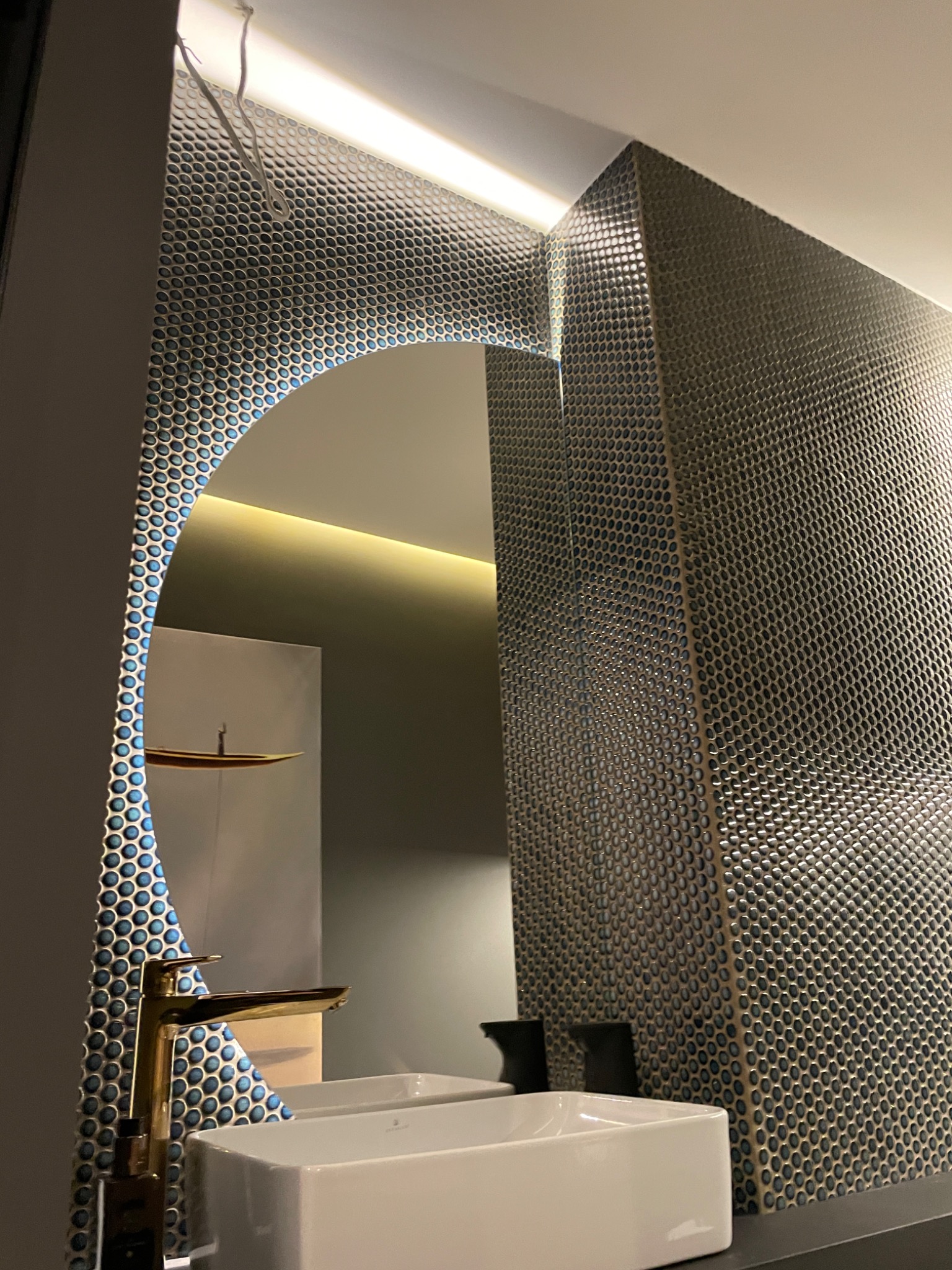 Nowoczesna łazienka z umywalką nablatową, złotą baterią i ścianą wykończoną mozaiką z drobnych okrągłych płytek, oświetlona listwą LED.