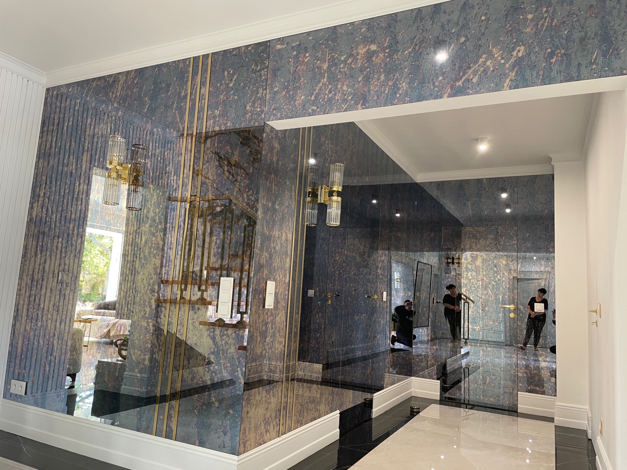 Lustrzane panele ścienne z dekoracyjnym wzorem w odcieniach granatu i złota, zamontowane w przestronnym holu z widocznymi pracownikami podczas montażu, odbijające schody i oświetlenie.