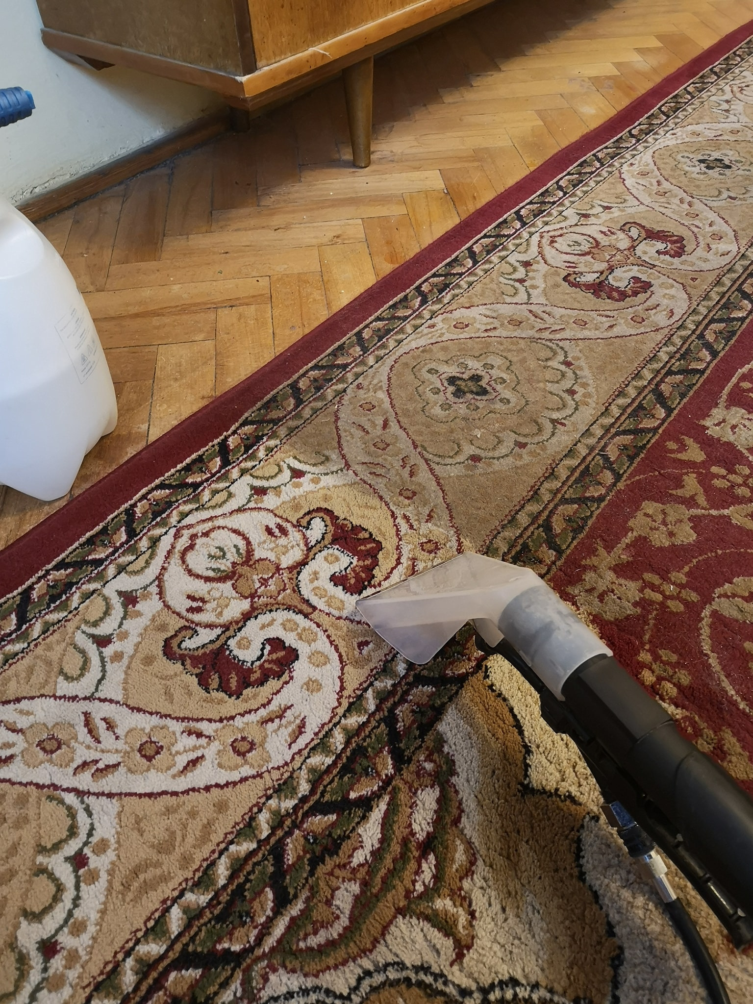 Czyszczenie wzorzystego dywanu metodą ekstrakcyjną, widoczna dysza odkurzacza piorącego i fragment drewnianej podłogi.