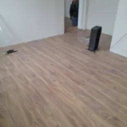 Świeżo ułożona, jasnobrązowa podłoga laminowana w pustym pokoju, widoczna listwa przypodłogowa, odkurzacz i grzejnik elektryczny.