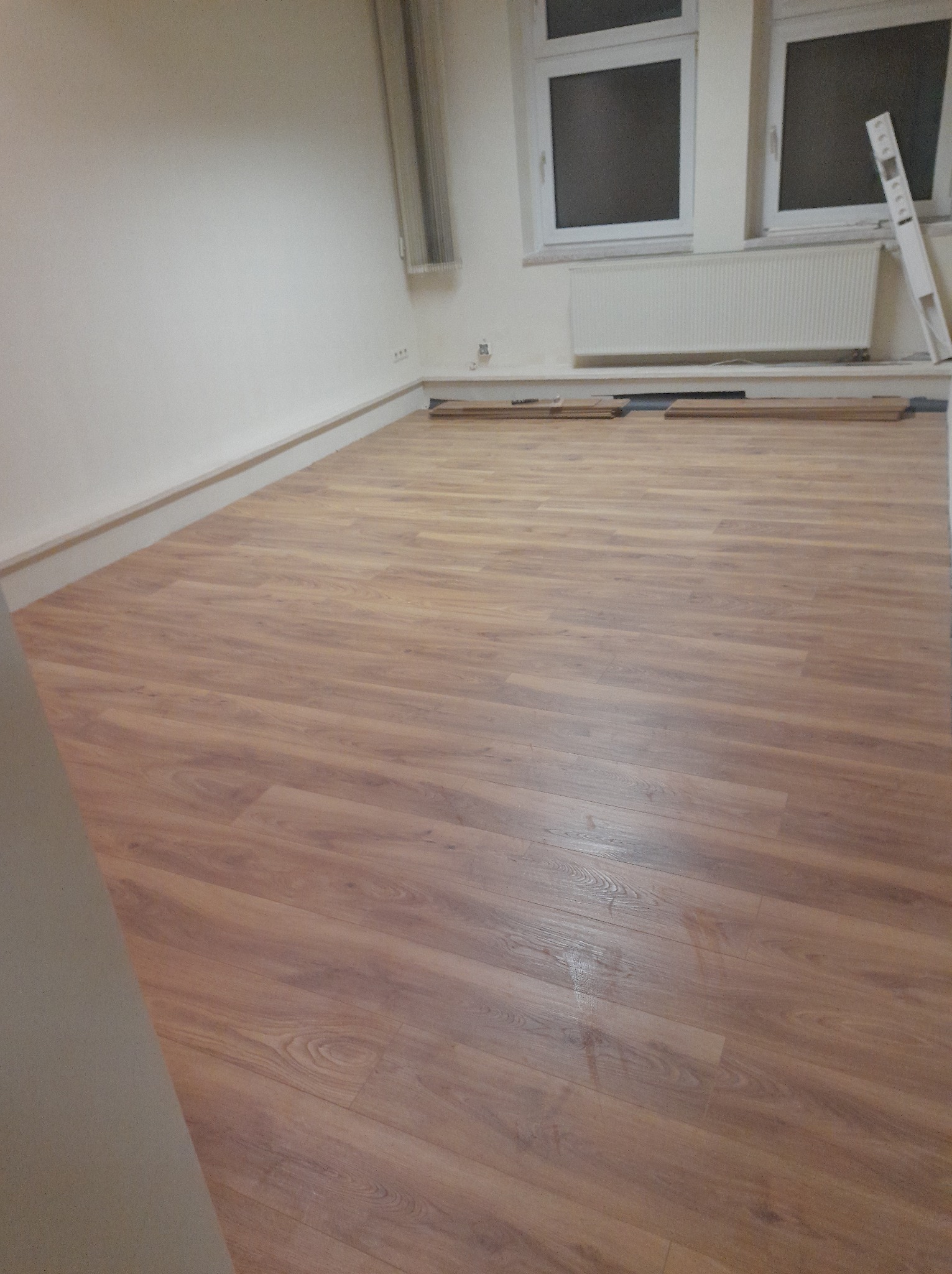 Ułożona podłoga z paneli imitujących drewno w jasnym pomieszczeniu z białymi ścianami, grzejnikiem i oknami, w tle leżą panele do dalszego montażu.
