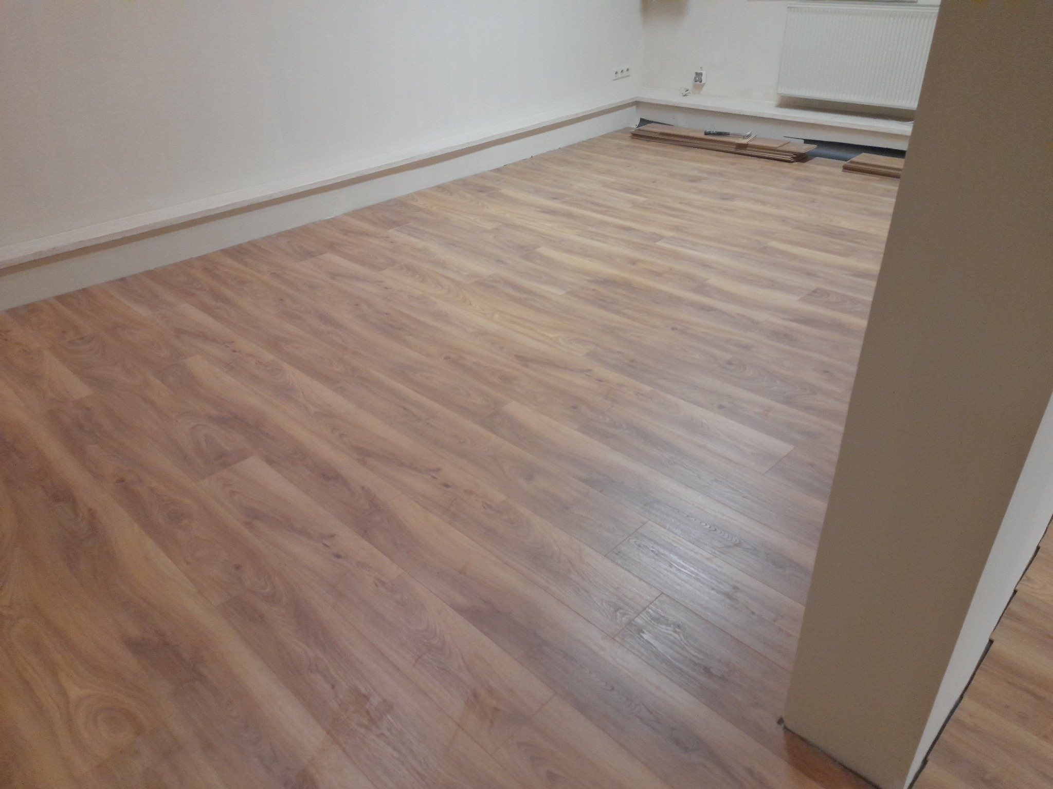 Nowo ułożona podłoga z paneli imitujących drewno w pustym pokoju, widoczne listwy przypodłogowe i stos paneli przy ścianie.