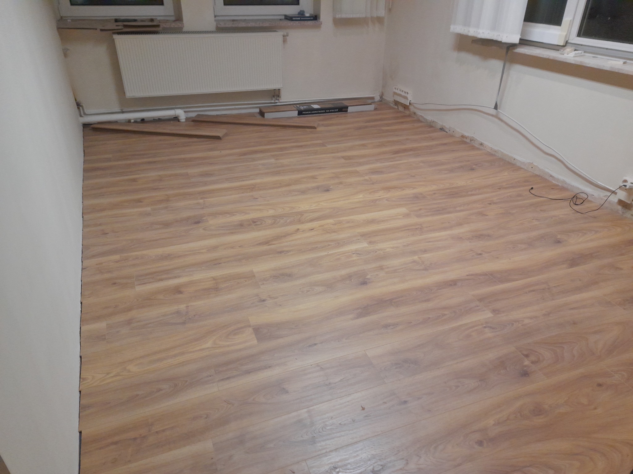 Wnętrze pokoju z ułożoną podłogą z paneli imitujących drewno, widoczne fragmenty nieułożonych paneli oraz opakowanie po panelach przy ścianie, grzejnik i fragment okna w tle.