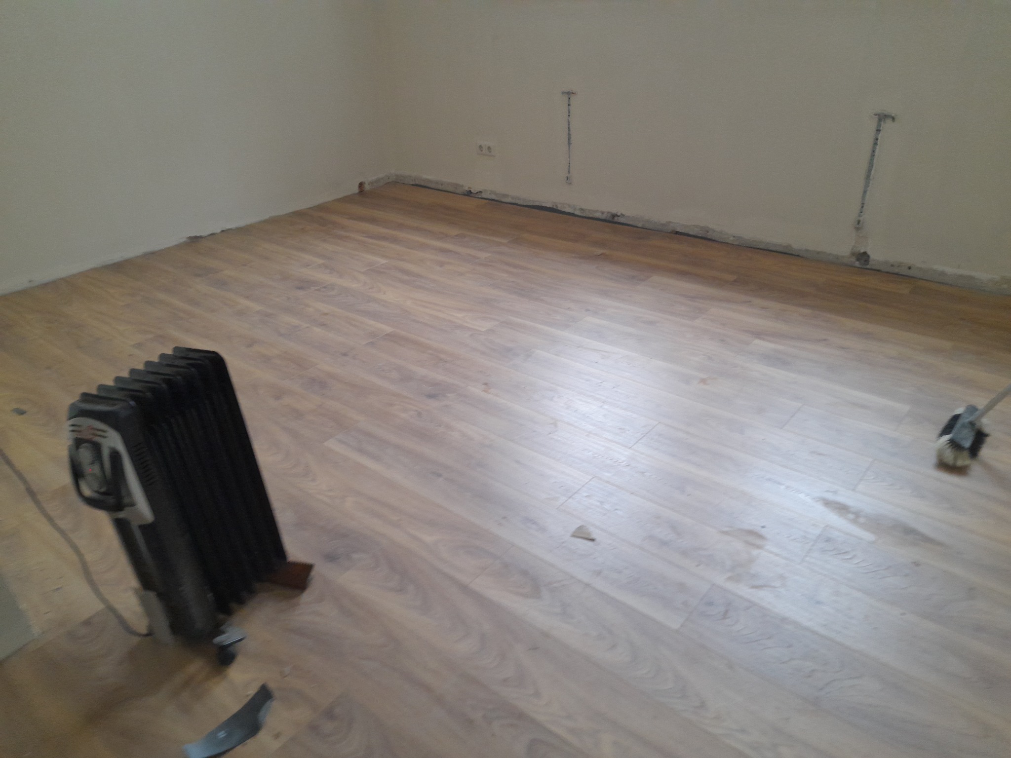 Nowo ułożona podłoga laminowana w pokoju, widoczny grzejnik elektryczny, szczotka do zamiatania i listwa przypodłogowa częściowo zamontowana.