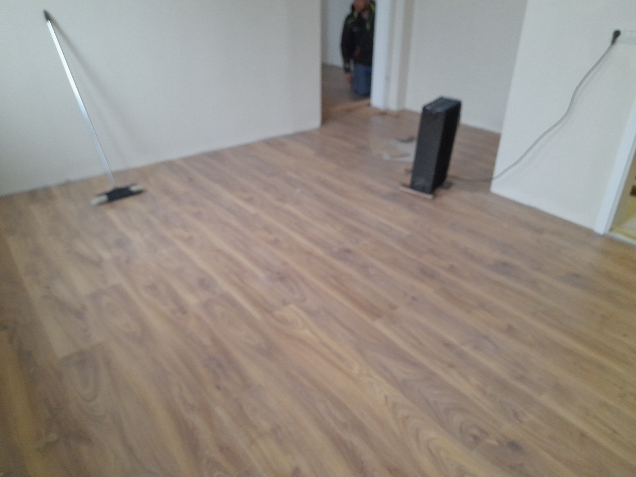 Świeżo ułożona, jasnobrązowa podłoga laminowana w pustym pokoju, widoczna listwa przypodłogowa, odkurzacz i grzejnik elektryczny.