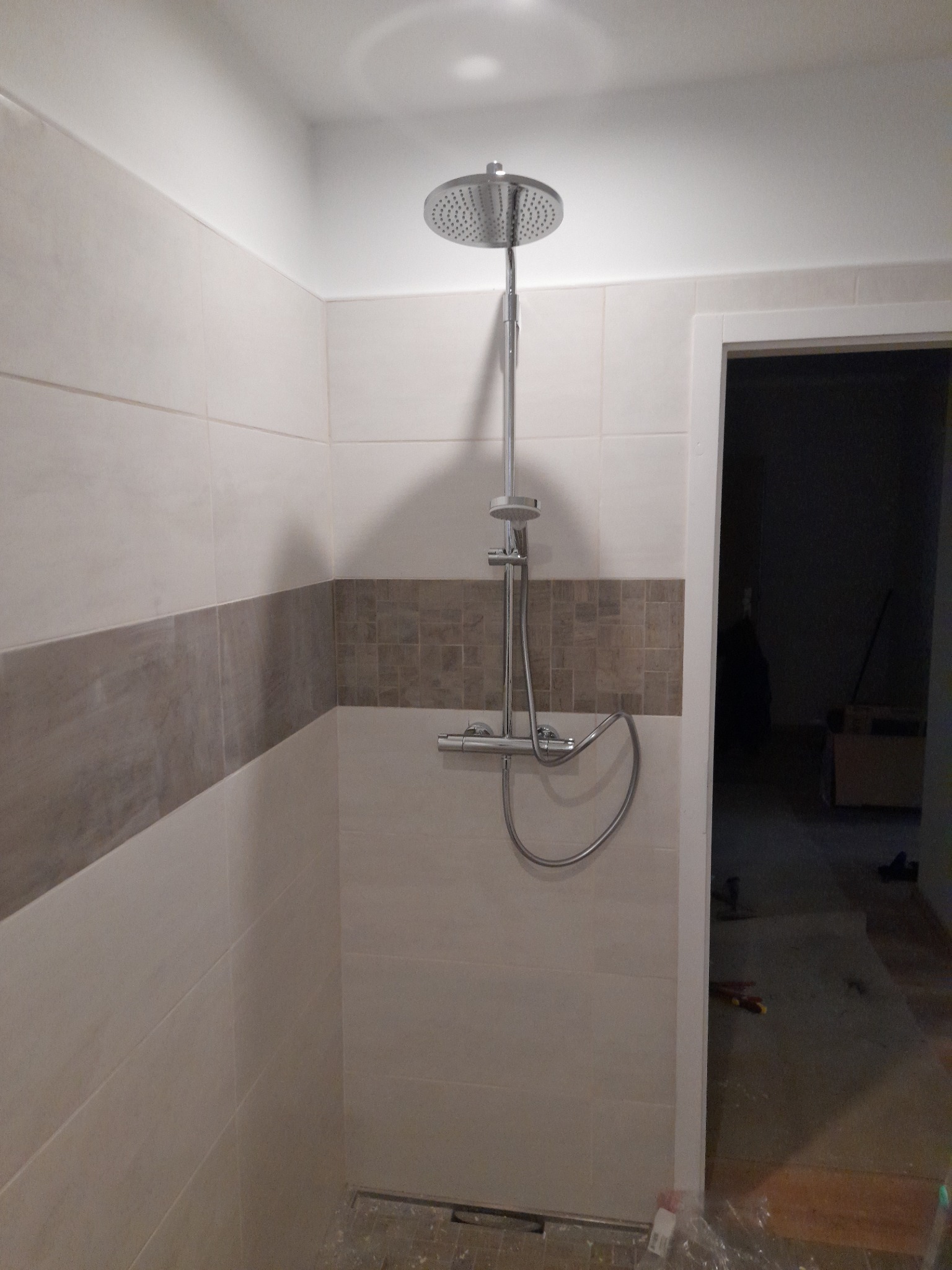 Nowoczesna kabina prysznicowa z deszczownicą i termostatem, wykończona beżowymi i brązowymi płytkami, widok na niedokończone pomieszczenie przez otwarte drzwi.