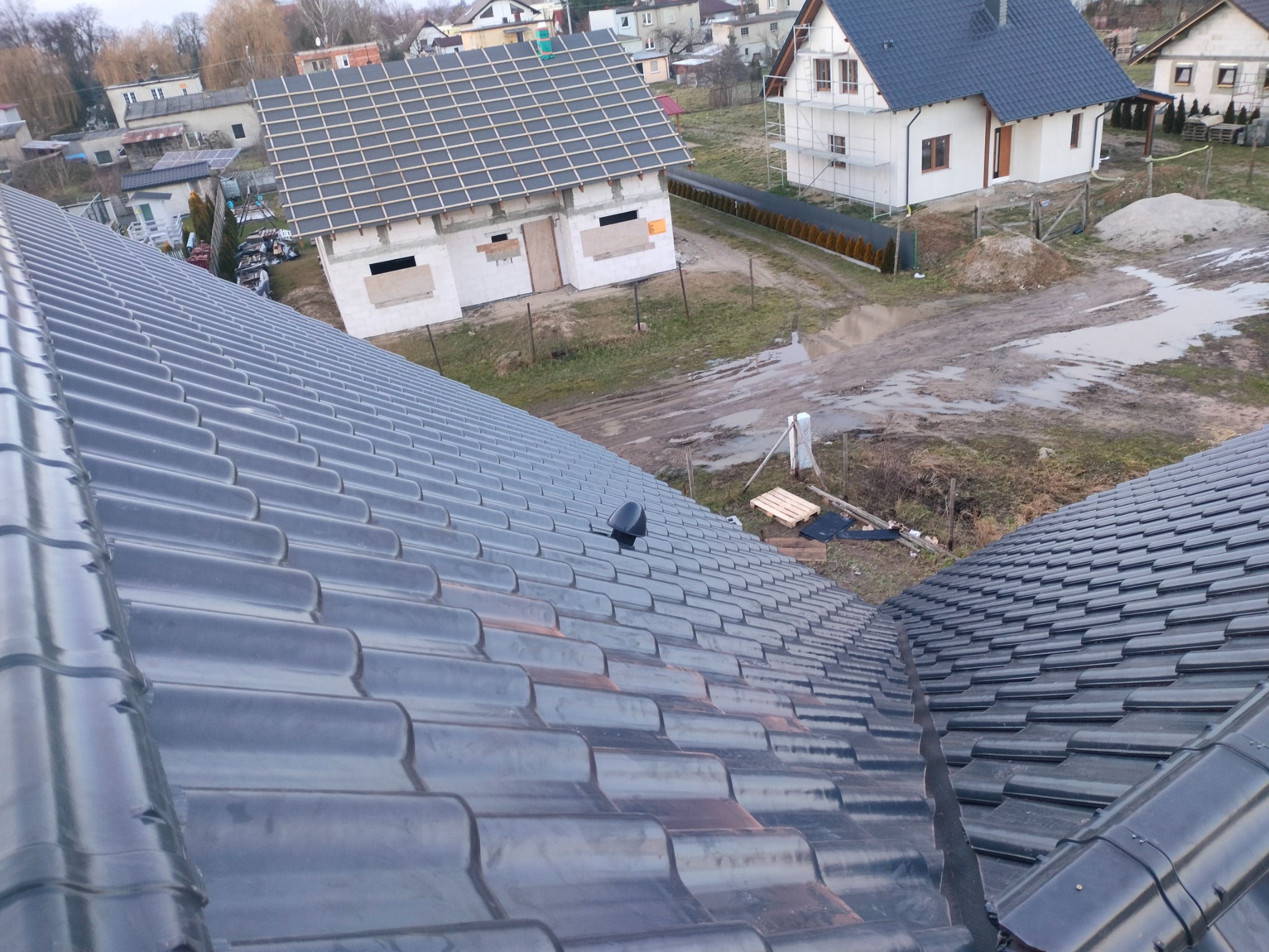 Widok z góry na błyszczące, grafitowe dachówki ceramiczne na nowym dachu, z widocznym kominkiem wentylacyjnym, w tle budynki w trakcie budowy i zabudowa jednorodzinna.