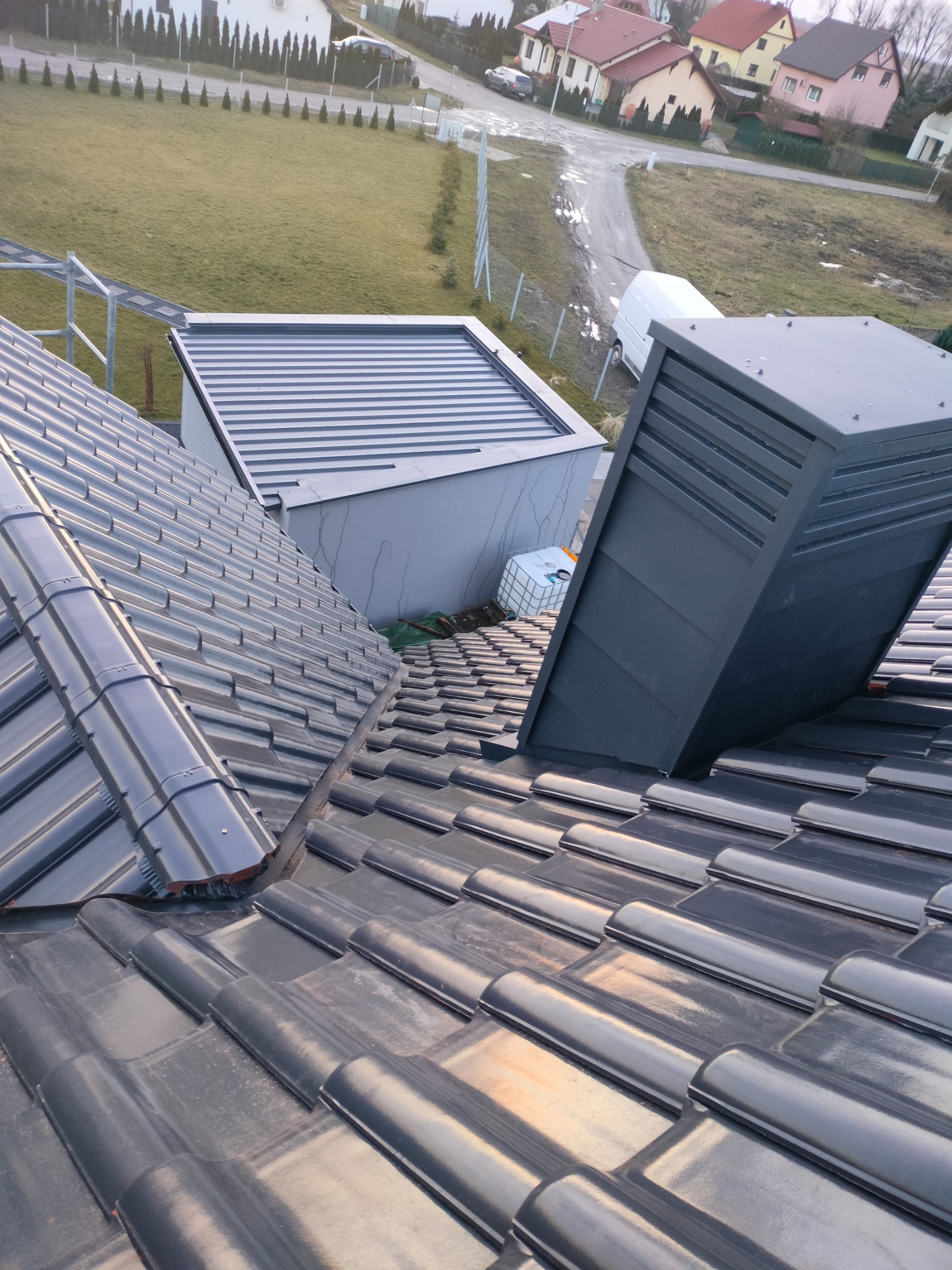 Nowy, ciemnoszary dach z dachówki ceramicznej, z widocznym kominem wentylacyjnym i fragmentem obróbki blacharskiej, na tle zabudowy jednorodzinnej.