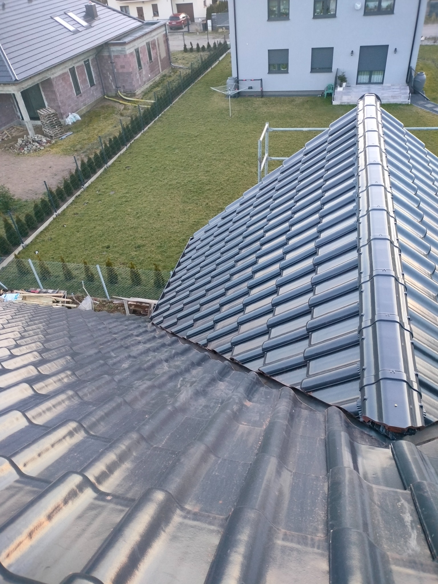 Widok z góry na fragment dachu z dwoma rodzajami dachówek: starsze, ciemniejsze i nowsze, błyszczące, z widocznym łączeniem oraz otoczeniem budynku i trawnikiem.