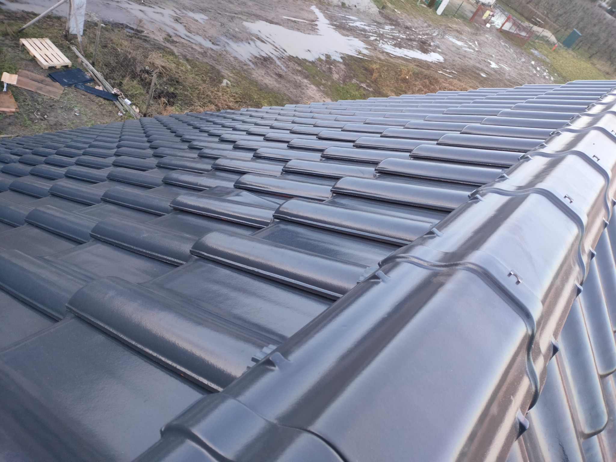 Nowo położona, błyszcząca dachówka ceramiczna w kolorze grafitowym, widziana z góry w Koronowie. W tle widoczny teren po deszczu.