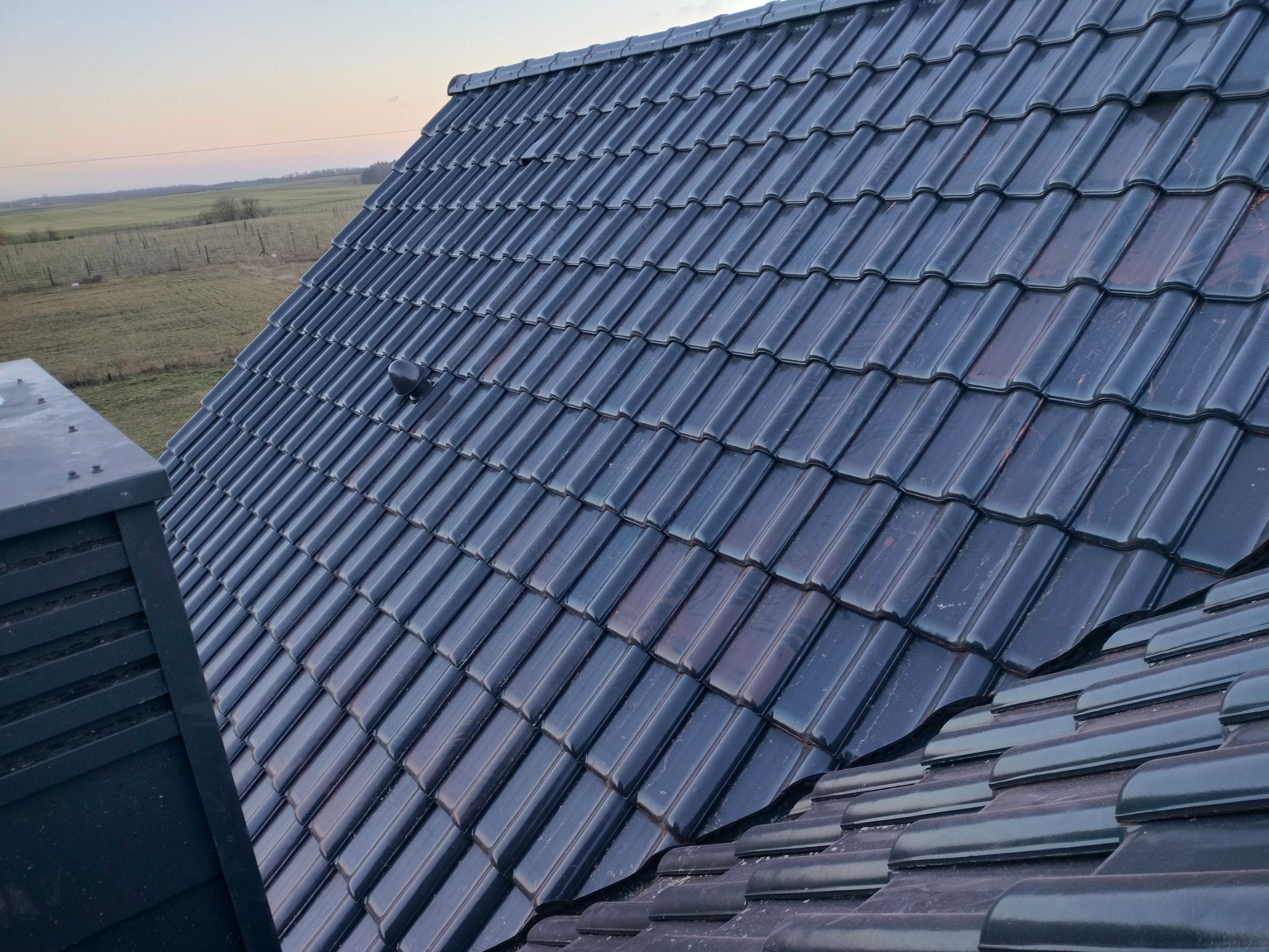 Błyszczące, ciemne dachówki ceramiczne ułożone na skośnym dachu domu, widok z góry, w tle krajobraz wiejski i zachodzące słońce.