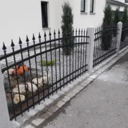 Czarna, metalowa barierka z ozdobnymi elementami i kamiennymi słupkami, oddzielająca ogród od chodnika, z widoczną przyczepką samochodową w tle.